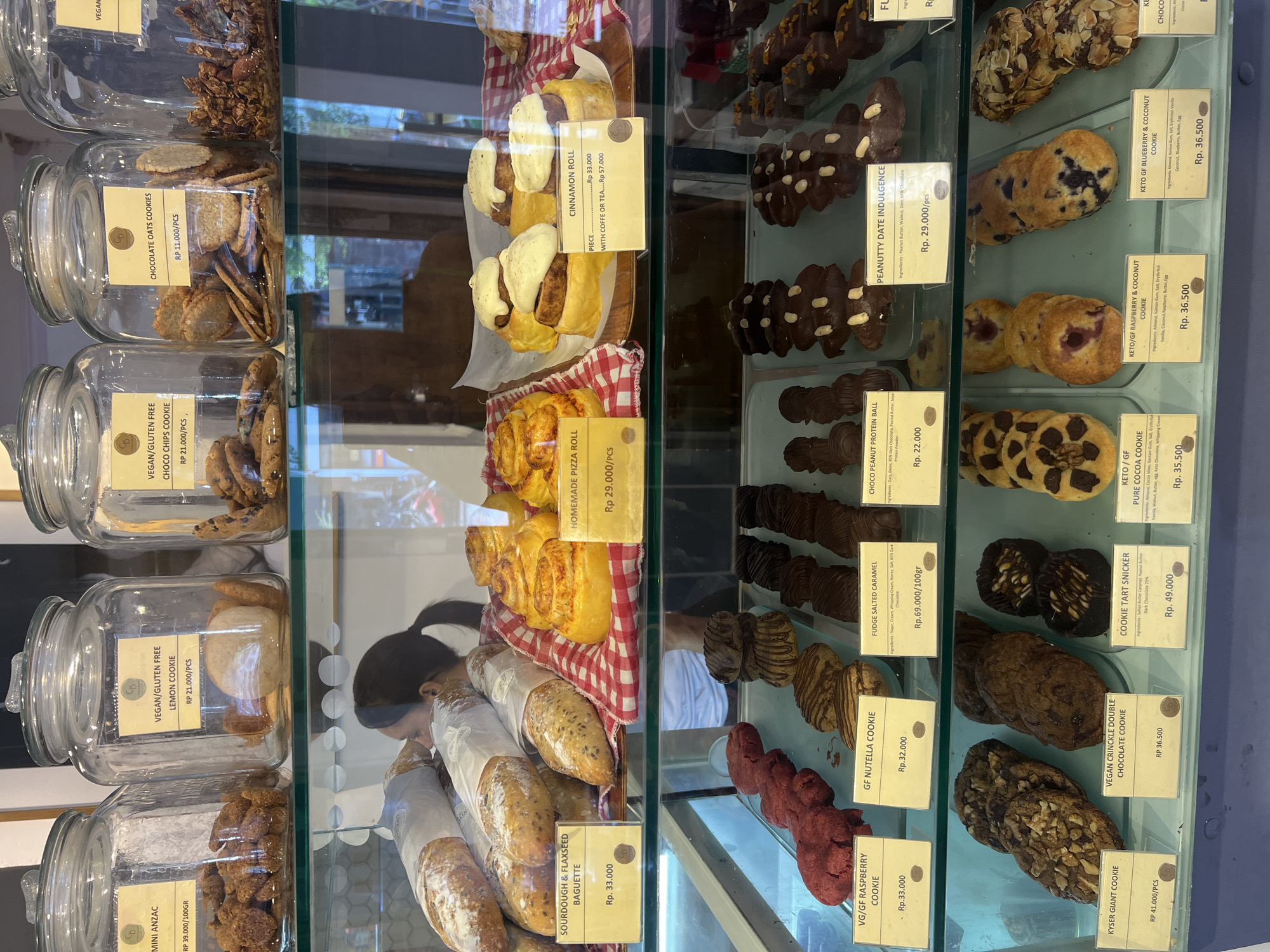 The image shows a display case in a bakery or cafe, likely located in North Kuta, Indonesia. The case is filled with a variety of baked goods, including cookies, rolls, bread, and other pastries. Glass jars on the top shelf hold individual cookies. The items are labeled with product names and prices in Indonesian Rupiah (RP). Some items are marked as vegan, gluten-free (GF), or keto-friendly. A woman is visible in the reflection in the glass.