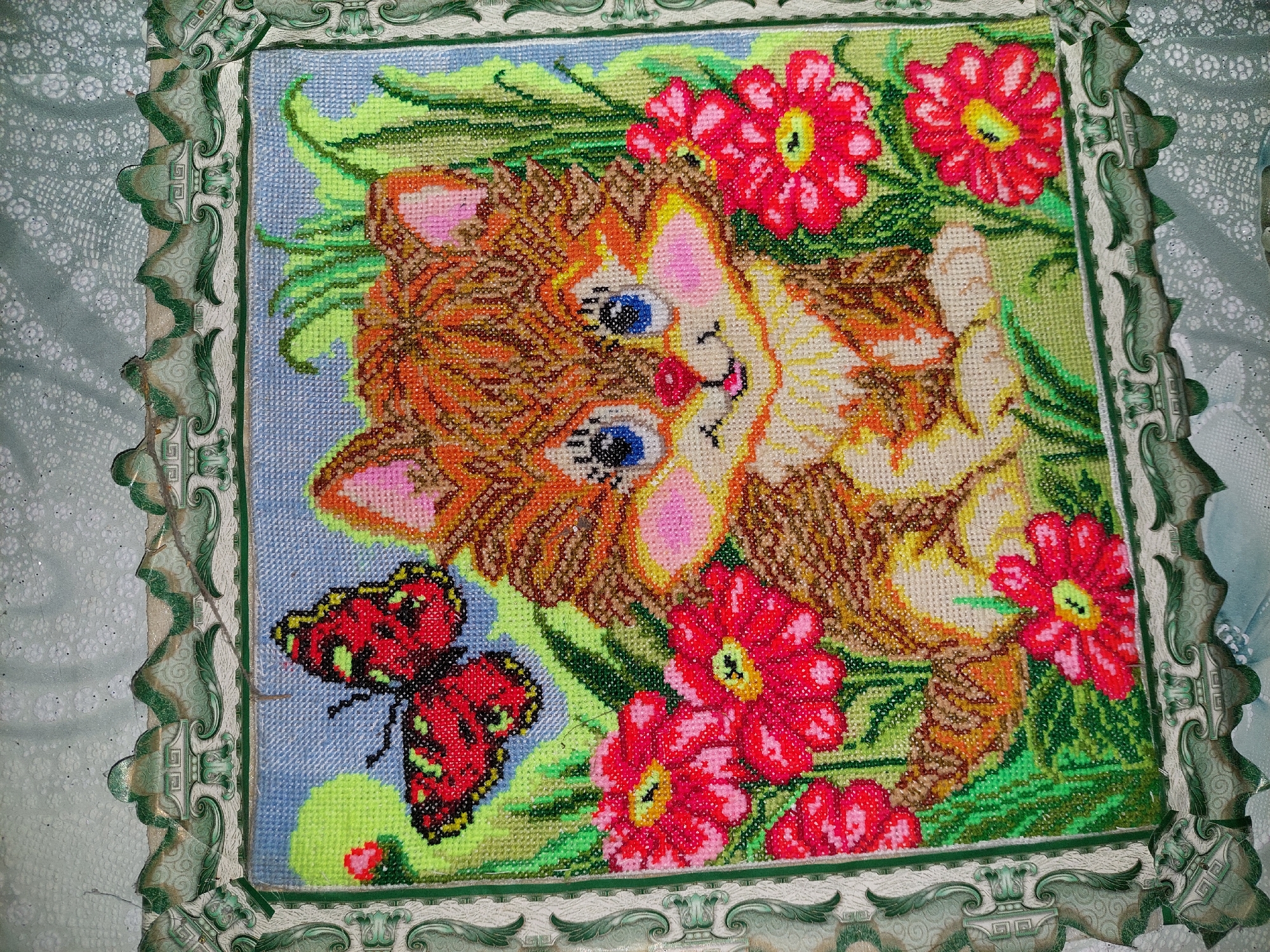 A beaded embroidery of a kitten and a butterfly is displayed in a decorative green and white frame. The kitten, depicted with orange and brown fur, blue eyes, and pink cheeks, sits amidst red flowers and green grass. A red and black butterfly hovers in the upper left corner. The background is a light blue sky. The artwork is likely a craft project, possibly intended for home decoration. The location is the village of Shpychyntsi, Ukraine.