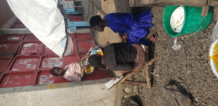 The scene depicts an outdoor setting in Buzove, Democratic Republic of the Congo. Three females are present: one stands leaning against a weathered red metal storefront door, wearing a light-colored, polka-dotted top and a patterned skirt; another is seated on a makeshift wooden stool with her back partially to the viewer, wearing a dark top and light brown bottom; the third is seated on the ground facing the stool, clad in a vibrant blue dress with red sandals visible near her feet. The two seated individuals appear to be conversing. Overhead, a large, worn white market umbrella provides shade. The ground consists of dark gravel and small stones. To the left, a concrete pillar is visible. In the foreground, a green bucket holds several white dishes, and a white bowl containing a yellow-orange substance rests on the ground.