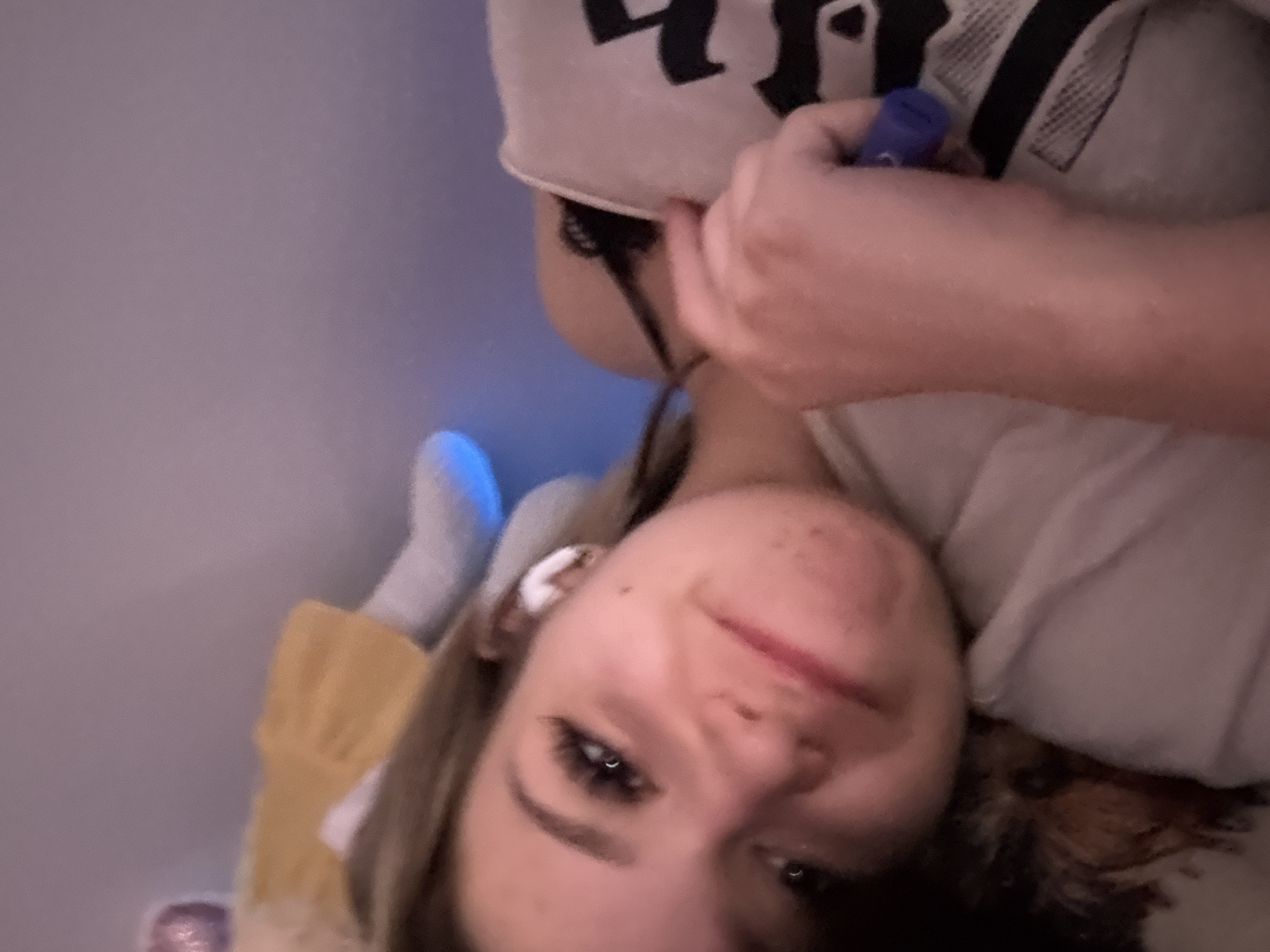 The image depicts a young individual, appearing to be female, positioned at an angle and looking directly towards the camera within an indoor, low-light setting. The subject has light brown hair, visible on the upper left. Facial features include prominent eyelashes, defined eyebrows, a small mole on the right cheek, and multiple small blemishes on the lower left facial area. A faint smile is present. The individual wears a light-colored top, possibly a t-shirt, with a partial black graphic design visible on the right chest area. A black strap, consistent with a bra strap, is visible over the right shoulder. A white wireless earbud is inserted into the right ear. The individual's left hand, partially visible, holds a small blue cylindrical object, consistent with a vaping device, near the chest. The background shows a plain wall illuminated by a blue ambient light source, originating from the lower right of the background. To the upper left, a portion of a light-colored stuffed doll, dressed in a yellow garment, is visible, along with a pink, possibly textured, spherical object. The scene is located in the city of Rezbartsi, Bulgaria.
