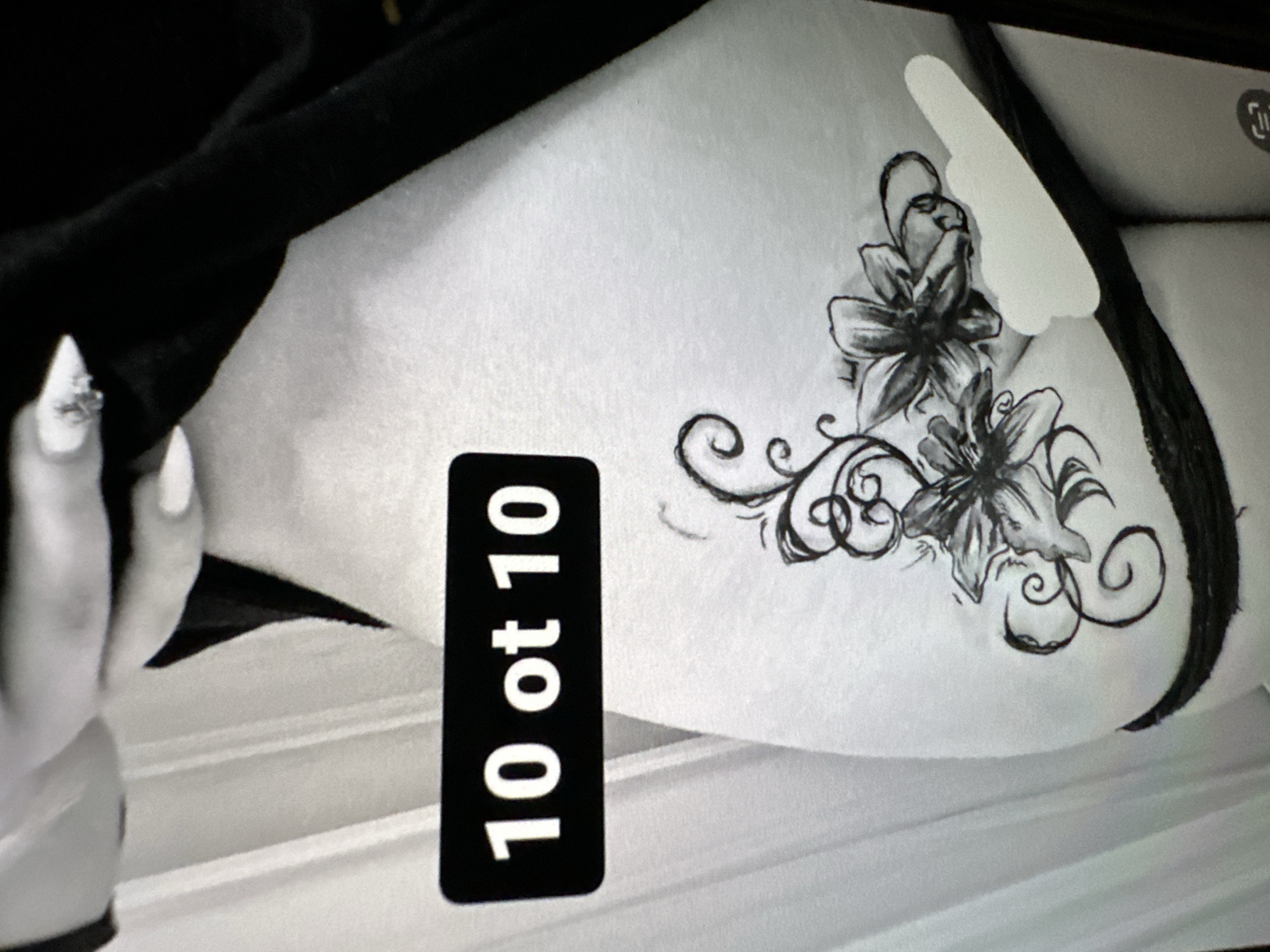 This black and white image features a close-up view of a person's hip and upper thigh, likely an indoor setting. A hand with a long, white, pointed fingernail, adorned with a ring or embellishment, is visible on the left, resting near a dark garment. The person is partially clad in a dark garment, possibly underwear or a bikini bottom, visible along the right side and peeking from under the hand. The central focus is an intricate floral tattoo on the hip, depicting multiple flowers with detailed shading and swirling vine-like patterns. A black rectangular digital overlay or sticker with white text "10 of 10" is prominently placed in the lower left. A white, irregular shape appears to be censoring a small portion of the image near the top right, and a faint icon (possibly a social media share button) is also visible in the upper right corner. The background consists of subtle horizontal lines, suggesting a textured surface such as bedding or a wall. The image itself appears to be a photograph of a screen, indicated by the visual quality. There are no visual cues to confirm the specified location of Varna, Bulgaria.