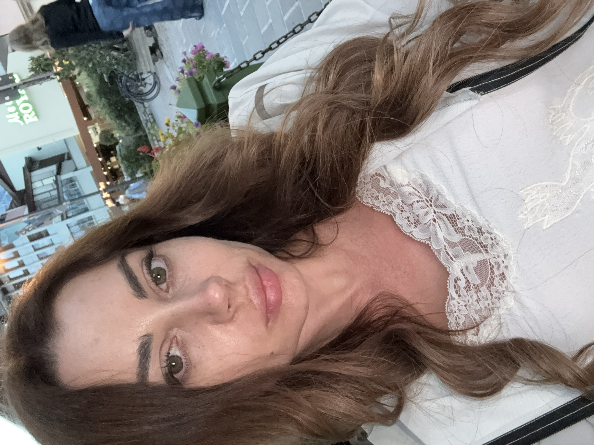 A woman with long, wavy brown hair is taking a selfie in an outdoor setting in Cortina d'Ampezzo, Italy. She is wearing a white top with lace trim. The background shows a building with signage that reads "Met. ROY." There are people and bicycles in the background, indicating a public area. The lighting suggests it is daytime.