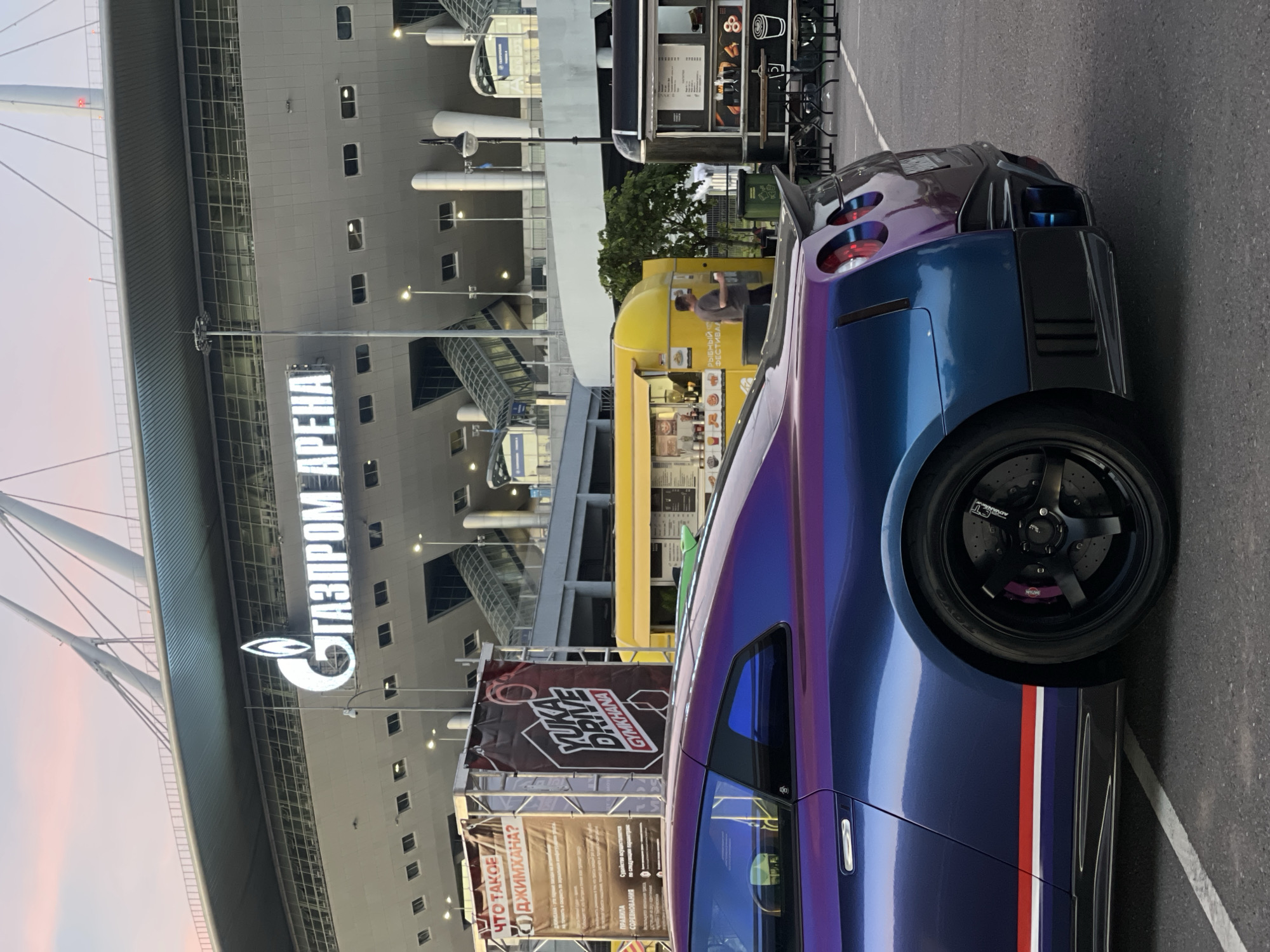 An eye-level, medium shot captures the rear of a metallic purple and blue Nissan GT-R parked in front of the Gazprom Arena stadium. The stadium's illuminated sign, "ГАЗПРОМ АРЕНА", is visible at the top of the image. To the left of the car, a banner displaying "YUKA DRIVE GYMKHANA" is mounted on a stand. In the background, yellow and dark-colored food trucks are parked, with a man walking between them. The scene is set in an outdoor paved area under a twilight sky.