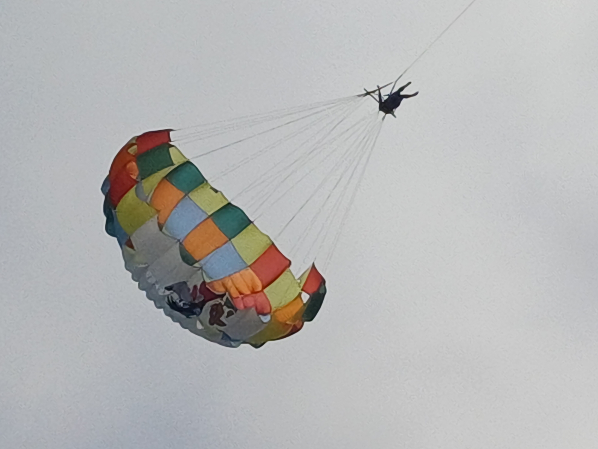 A parasailer is captured in mid-air against a pale, overcast sky, likely over the waters near Sochi, Russia. The parasail is a colorful patchwork of orange, yellow, green, blue, and red squares, with a faded image of what appears to be a cartoon character or graphic visible on the upper portion. The parasailer, dressed in dark clothing, is suspended beneath the canopy by thin white lines. The person's legs are visible, and they appear to be in a relaxed posture. The overall scene suggests a recreational activity taking place outdoors.