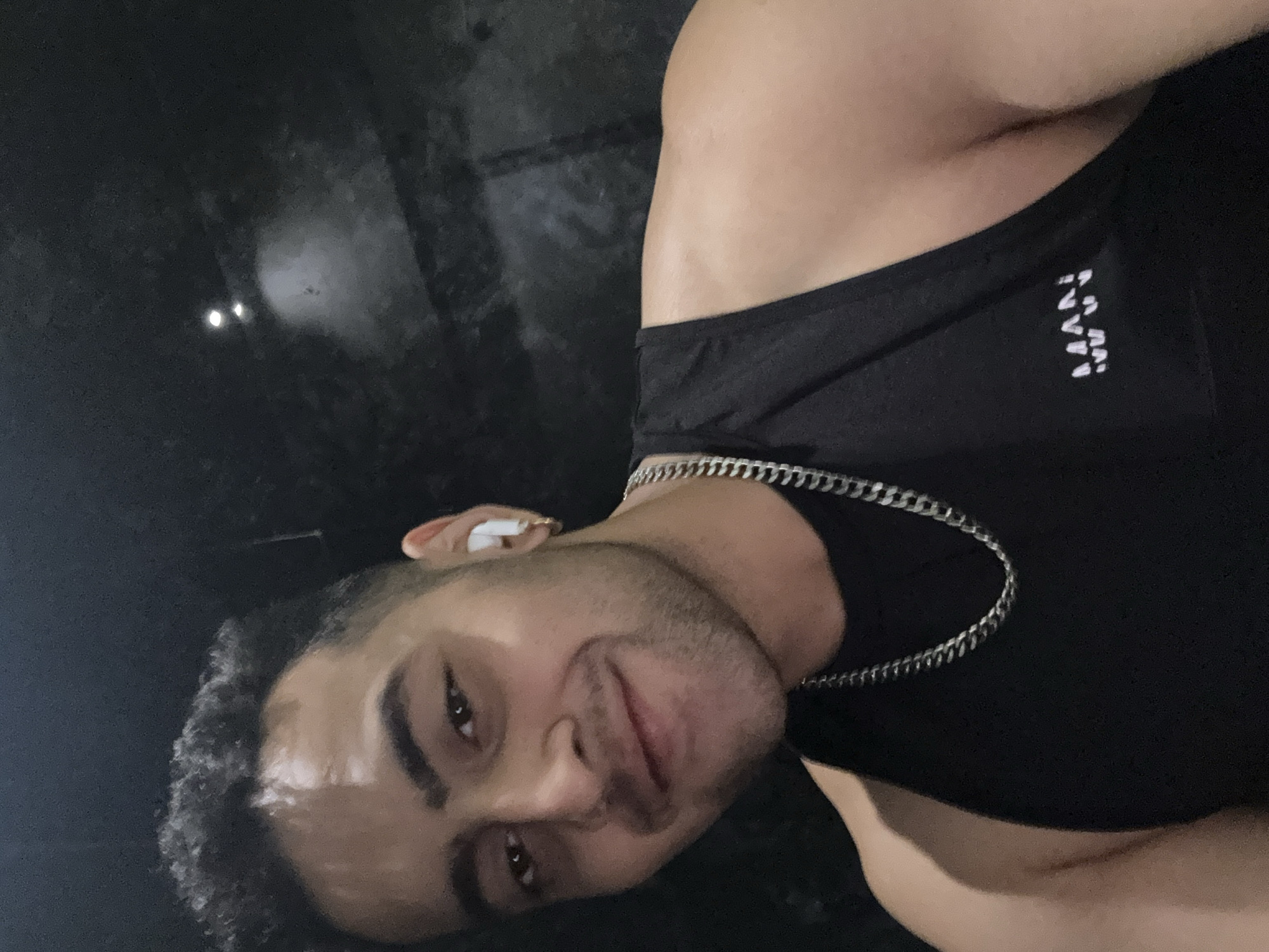 A close-up, low-angle shot features a man with dark hair and eyes, fair skin, and light stubble on his face. He is wearing a black tank top with the word "MAN" vertically printed in white on the right shoulder. A silver chain necklace is around his neck. He has a wireless earbud in his right ear. The background is dark and indistinct, with two blurry spots of light visible. It is not possible to determine the location as Kolkata, India from the provided image.