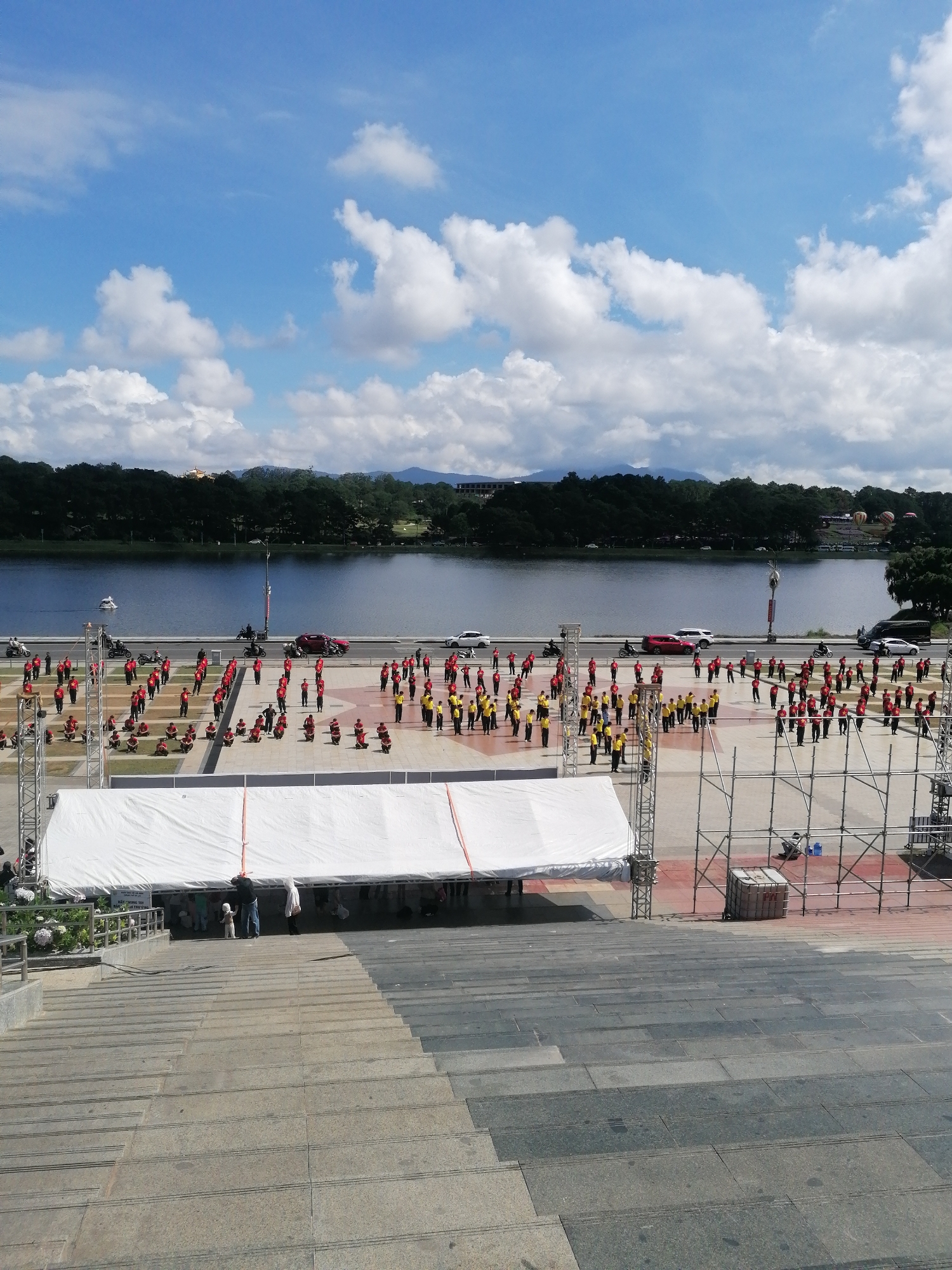 In Da Lat, Vietnam, an outdoor event is underway near Xuan Huong Lake. A large group of people, many dressed in red and yellow, are assembled on a paved area. Some are standing in formation, while others appear to be seated. A white tent structure, supported by scaffolding, is set up in the foreground. Beyond the paved area, the calm waters of Xuan Huong Lake are visible, bordered by a tree-lined shore and distant mountains. Vehicles, including cars and motorcycles, are present on a road running alongside the lake. The sky is partly cloudy with patches of blue.