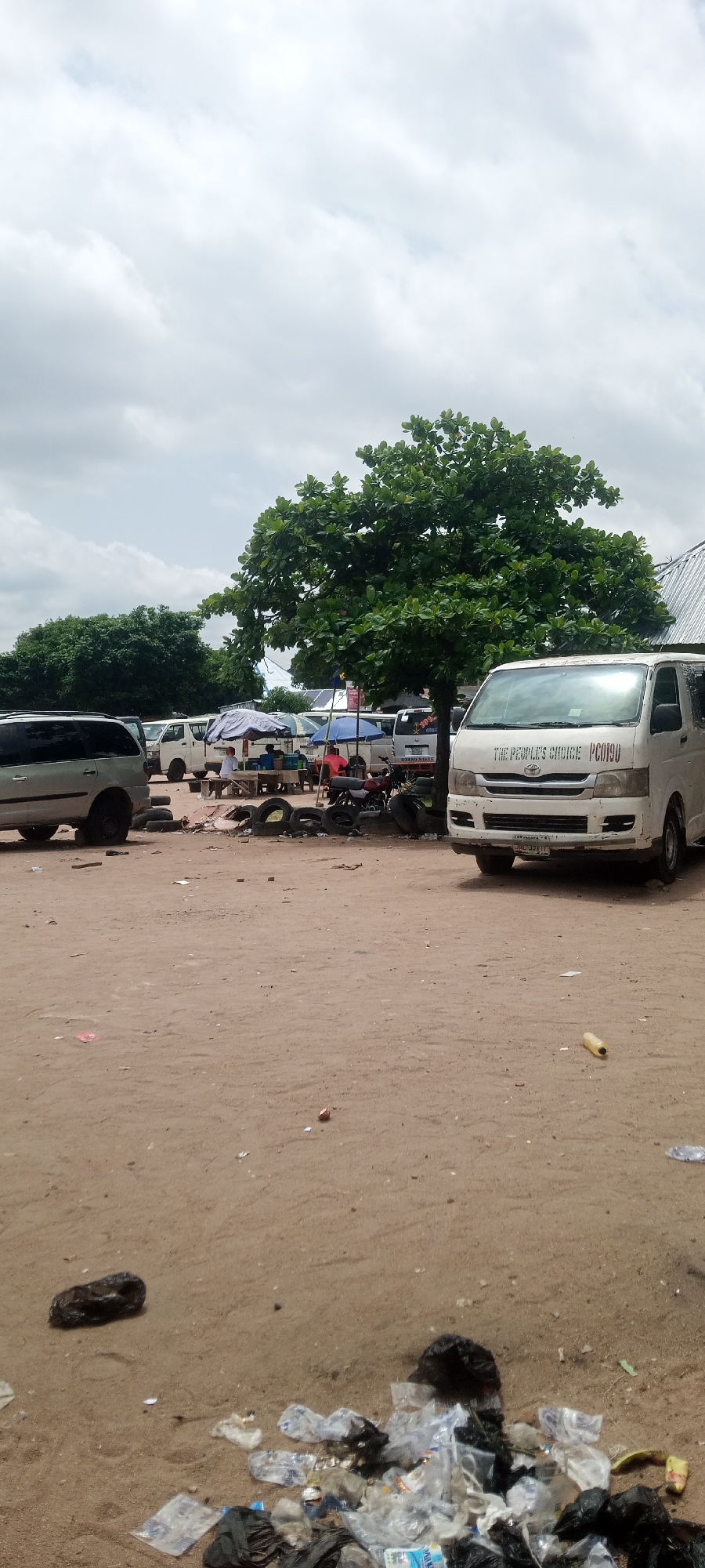 The image shows an outdoor scene in Jalingo, Nigeria, likely a bus or taxi park area. A white van with "THE PEOPLE'S CHOICE PCO190" written on its front is parked on the right side of the frame. Several other white vans and a silver SUV are visible in the background. There are people sitting under umbrellas, likely waiting for transportation or selling goods. Tires are scattered around the area, some appearing used as seating or as part of the ground. The ground is sandy and dusty, with visible trash and debris, including plastic bottles and black garbage bags, particularly in the foreground. A large, leafy tree stands prominently in the mid-ground. The sky is overcast with clouds.