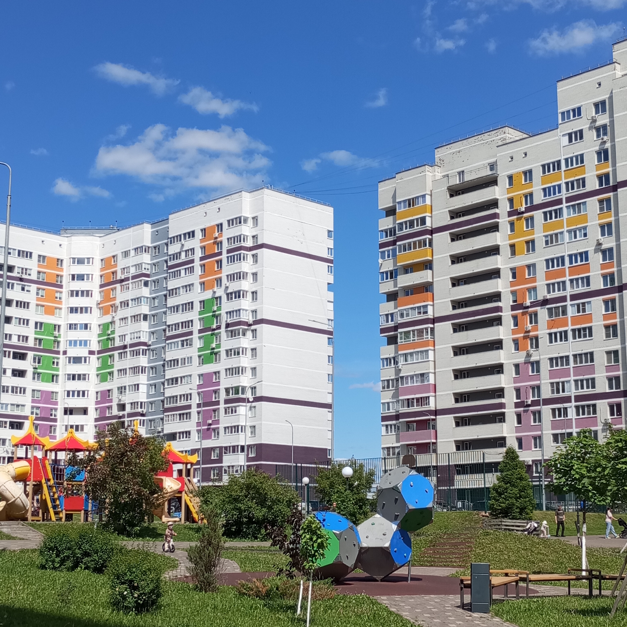 The image displays a residential park area with modern apartment buildings under a clear blue sky with scattered white clouds, located in the city of Pervomaysky, Russia.
Two multi-story apartment buildings dominate the background. The building on the left features a white facade with colored rectangular sections in orange, green, and purple around windows and balconies. The building on the right presents a light grey facade accented with yellow, orange, and purple panels on its balconies and window surrounds. Numerous windows and balconies are visible on both structures, with several external air conditioning units mounted on the right building.
The foreground and middle ground comprise a well-maintained park. On the left, a large, colorful children's playground structure includes yellow slides, red roofs, and climbing elements on a light-colored ground surface. On the right, a contemporary climbing sculpture, assembled from interconnected polyhedral units in blue, green, and grey, sits on a brown rubberized safety surface. Green grass covers the ground, punctuated by various bushes and small trees. Paved pathways of light grey pavers wind through the landscape.
Several individuals are present. A small child, wearing a helmet, rides a balance bike on a paved path near the left playground. On an elevated paved path to the right, a person in a dark shirt stands near a wooden bench. Another person sits on the bench, with a small white dog positioned next to them. Further along the uphill path, two individuals walk, one pushing a stroller.
Additional objects include a metal lamppost partially visible on the far left, a tall dark metal fence behind the geometric climbing structure, and a dark grey trash receptacle situated next to a wooden park bench in the lower right foreground. Bright sunlight illuminates the scene, creating distinct shadows.