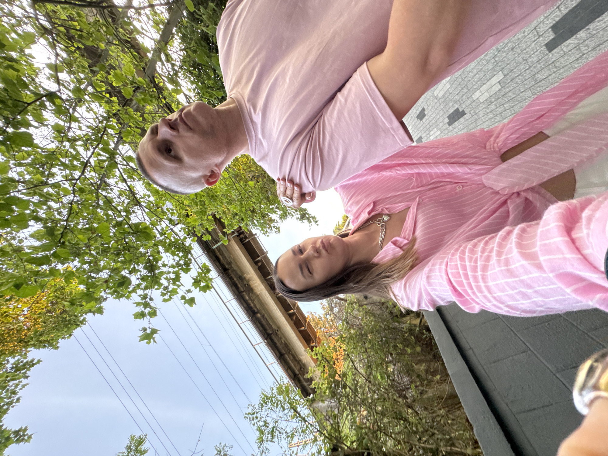This is a selfie taken outdoors with a slightly low-angle perspective. In the foreground, a man and a woman are visible. The woman is on the left, looking at the camera with a playful expression, winking. She is wearing a pink and white striped button-down shirt tied at the waist and a silver necklace with a heart pendant. The man is on the right, looking upwards and to the right, with his right shoulder slightly hunched. He is wearing a pink t-shirt. The woman's left hand is on the man's right shoulder.
Behind them, a dark grey, textured wall runs along the bottom of the frame. Beyond the wall, there is lush green foliage and trees. An elevated concrete structure, possibly a bridge or railway overpass, is visible in the background, spanning across the scene. Power lines are also visible against the sky. The ground appears to be paved with grey and white rectangular stones.
The lighting suggests it is daytime, with some shadows cast by the foliage. The overall impression is a casual, candid moment captured between two people outdoors. The location is the city of Sochi, Russia.