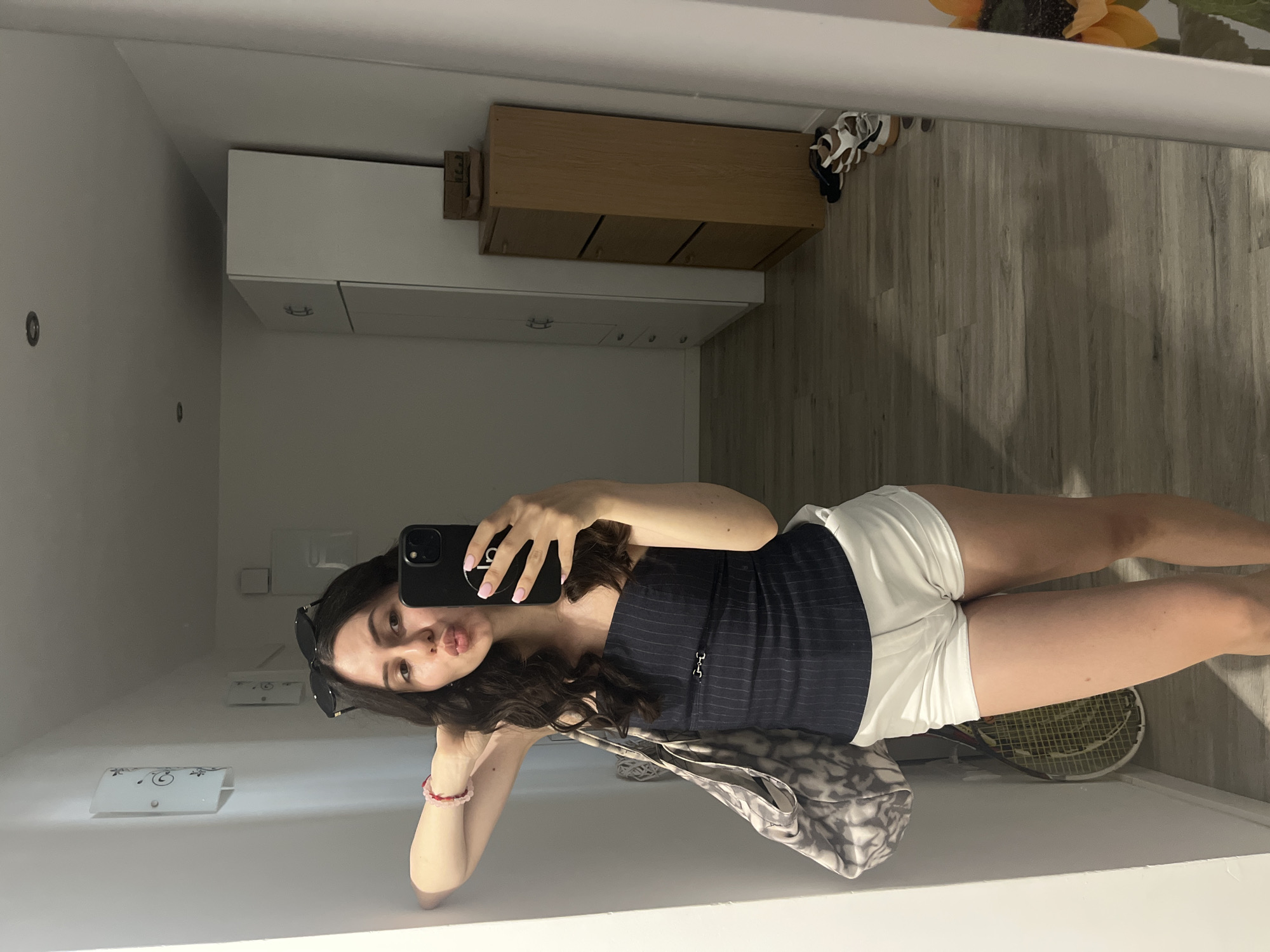 The image shows a young woman taking a mirror selfie. She is wearing a dark, pinstriped tank top and white shorts. She has long, dark, wavy hair and is wearing sunglasses perched on her head. She is leaning against a white surface and has a bag with a leopard print draped over her arm. Her left hand is holding her phone, and her right arm is bent, resting her head.
In the background, reflected in the mirror, are white cabinets with wooden accents, a wooden shelf unit with three drawers, and a glimpse of a sunflower and a tennis racket on the floor. The floor appears to be made of light-colored wood-look laminate.
The scene appears to be indoors, likely in a home or a dressing room, given the presence of furniture and storage. The lighting suggests it could be daytime, but there are no explicit indicators of the time of day or weather. The woman's pose and expression, with her lips puckered, suggest a playful or casual mood. No visible text is present in the image. The provided location context of Palma de Mallorca, Spain, adds a geographic element to the scene, though no specific local cues are visible in the image itself.