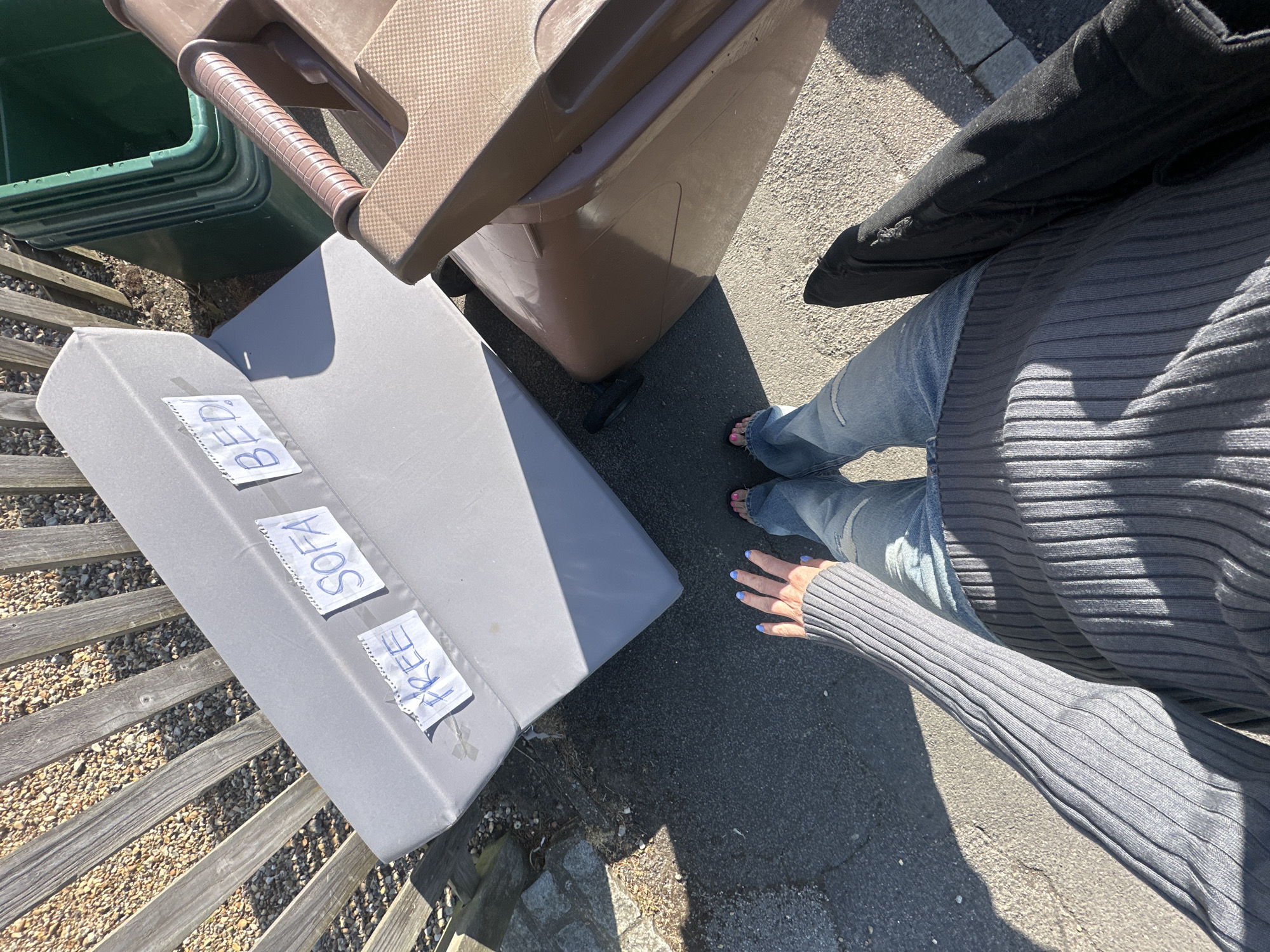 The image depicts an outdoor scene, likely in a residential area of Tunbridge Wells, United Kingdom. A woman, dressed in blue jeans and a gray ribbed top, is partially visible from the waist down. Next to her, on an asphalt surface and adjacent to a wooden fence, is a light gray rectangular object labeled "FREE SOFA BED". The object is placed near two refuse bins: a brown wheelie bin and a stack of green bins. A black jacket is partly visible between the bins and the woman. The scene is well-lit by sunlight.