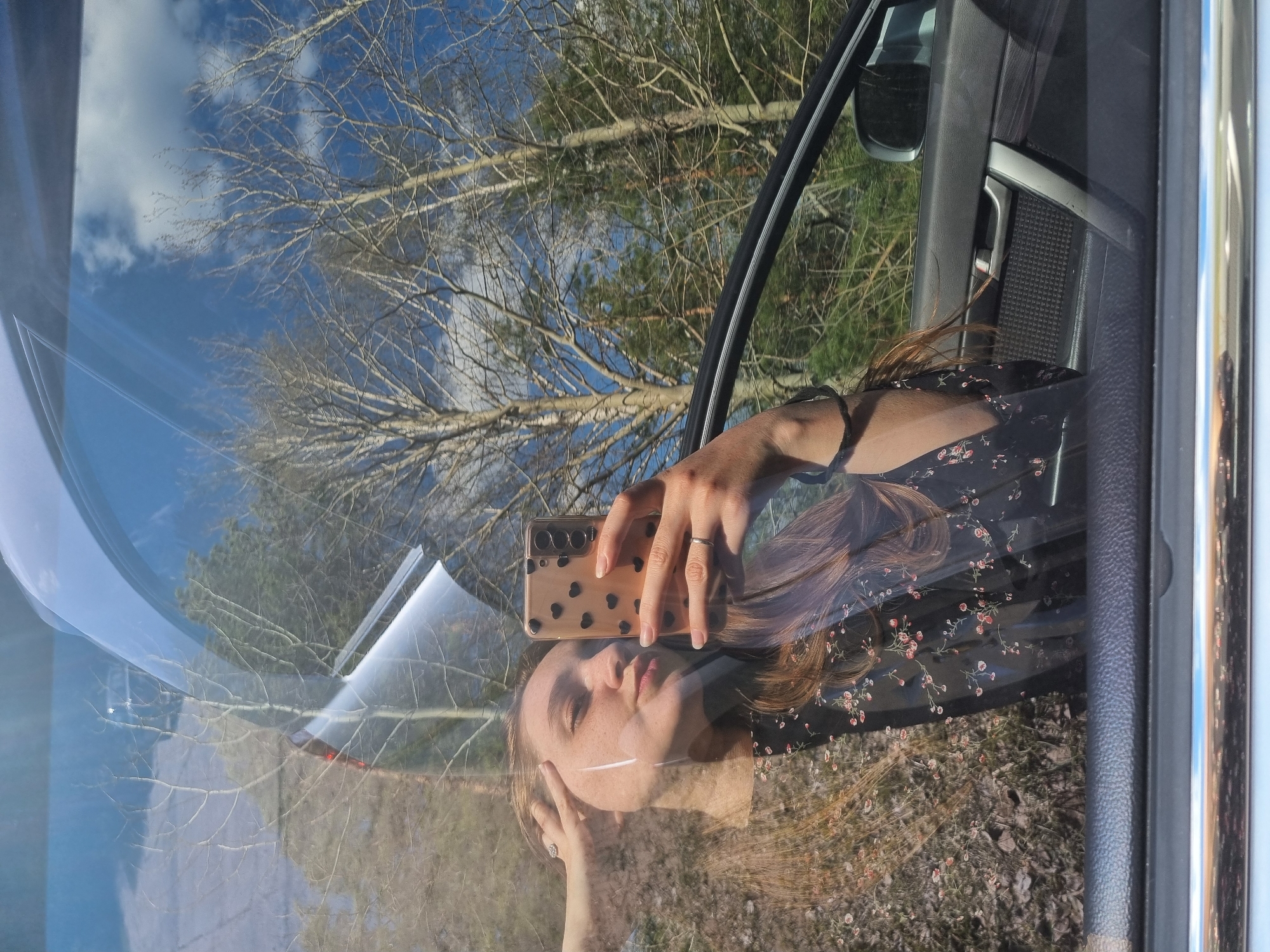 A woman is taking a selfie in a car. Her face and hand holding a cell phone are reflected in the windshield. The reflection in the windshield also shows bare trees and a blue sky with clouds. The car's interior, including the door panel, is visible. The location is Unknown, Unknown.