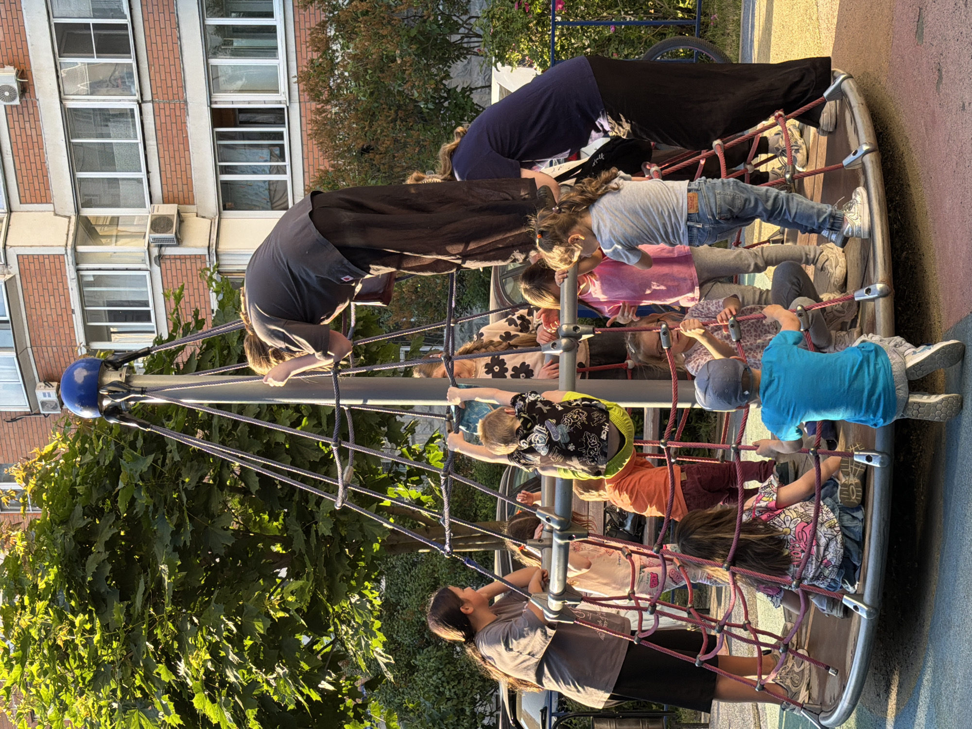 This image depicts a lively scene at a public playground, likely located in Khimki, Russia. The central focus is a large, circular climbing structure made of ropes and metal, resembling a net-like merry-go-round.
Numerous children of various ages are actively engaged with the climbing frame. Some are seen climbing the red ropes, while others are standing or sitting within the circular base of the structure. Notable children include one in a light blue t-shirt and blue jeans, another in a bright blue t-shirt and a blue baseball cap, and several others in casual, brightly colored or patterned clothing.
Several adults are also present, appearing to supervise or assist the children. Two adults are prominently featured leaning over the climbing structure: one in a dark blue long-sleeved top and black pants, and another in a dark grey t-shirt and black pants. The overall mood suggests a pleasant, active, and social outdoor gathering.
The scene is set outdoors on what appears to be a sunny late afternoon or early evening, evidenced by the warm, golden light illuminating the foliage and casting subtle shadows. The weather is clear. In the background, a multi-story brick apartment building with numerous windows and visible air conditioning units provides an urban context. Lush green trees with dense foliage are scattered around the playground area, suggesting a summer or early autumn season. The playground ground is paved with a colorful surface, featuring shades of red and blue. A portion of a bicycle wheel is visible in the upper right. No visible text is discernible in the image.