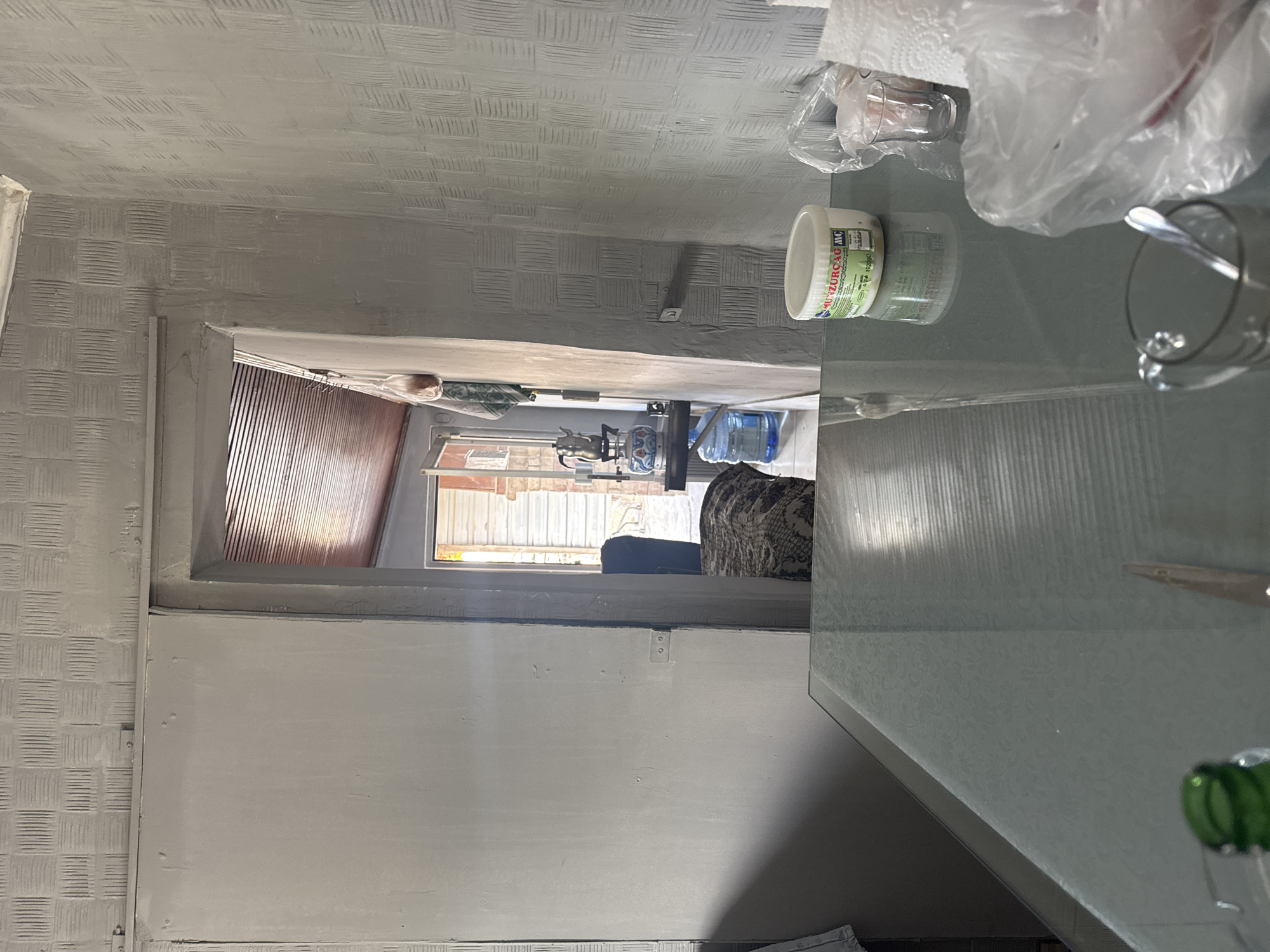 The image depicts an interior scene, likely a kitchen or dining area, with a glass table in the foreground. On the table, there are several items including a container of "MUNZURÇAM" cheese, a small glass, a roll of paper towels, and a plastic bag containing food items. A green bottle is partially visible on the left. The background shows a doorway leading to another room, where a traditional Turkish tea pot is visible on a shelf, along with a large water jug. The walls have a textured pattern. The scene appears to be an indoor residential space in Gebze, Turkey. No individuals are present in the image.