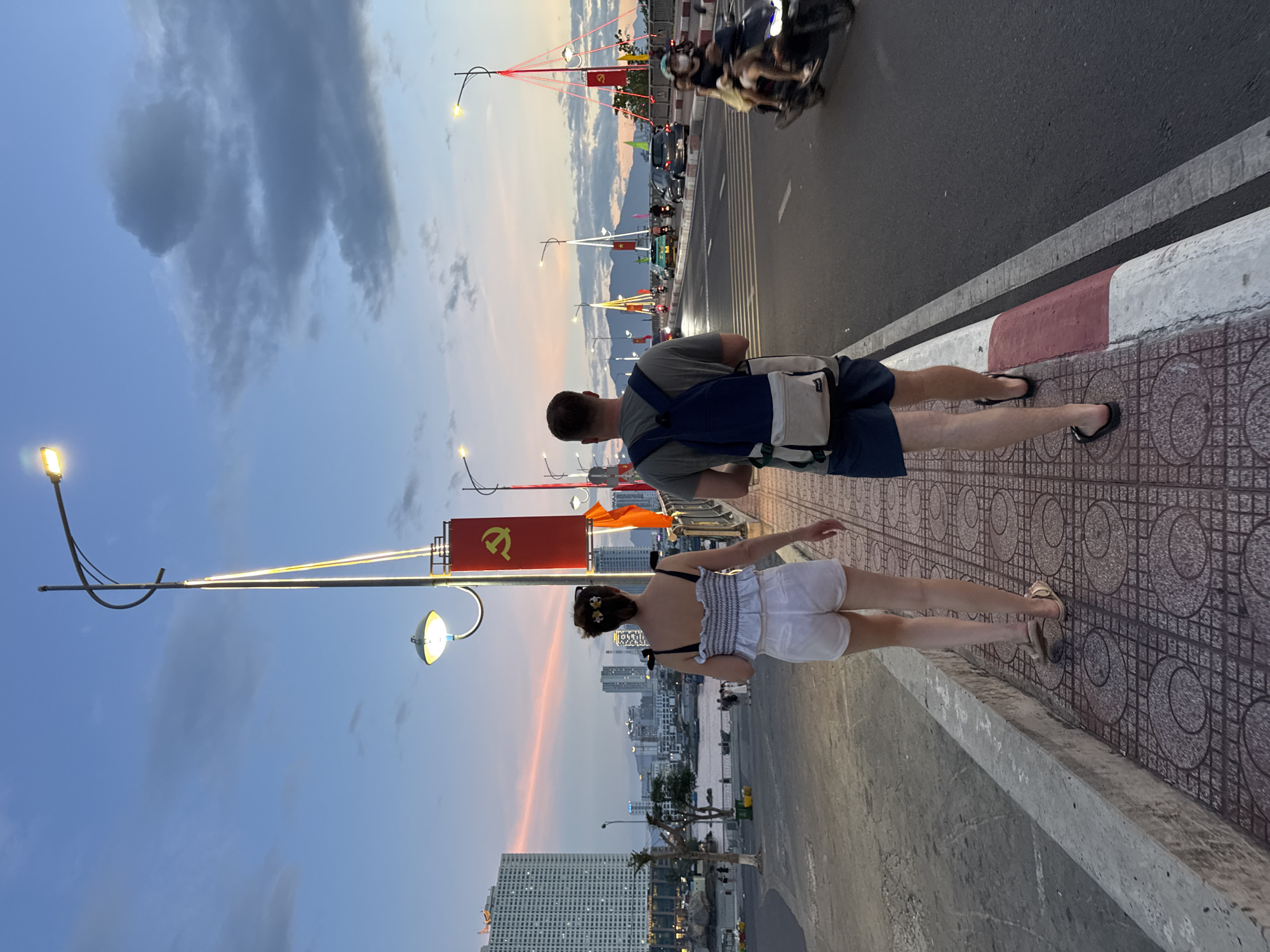 The image captures a sunset scene on a bridge in Nha Trang, Vietnam, likely Nha Trang Bay Bridge or Tran Phu Bridge, given the city skyline and coastal setting. A woman and a man are walking away from the camera on the pedestrian walkway. The woman wears a white ruffled top and white shorts with sandals. The man, with a backpack, wears a grey t-shirt and blue shorts with flip-flops. They are walking towards the right side of the frame.
The bridge is adorned with lampposts, some illuminated, and festive red banners with yellow symbols, possibly related to a national holiday or event. The sky displays a gradient of sunset colors, from soft pinks and oranges to a dusky blue, with scattered clouds.
Across the road, traffic flows with cars and motorcycles. One motorcycle, carrying two people, is in motion on the right side of the roadway. In the background, the cityscape of Nha Trang is visible, featuring tall buildings and structures along the coastline. The overall atmosphere suggests a pleasant evening stroll in a vibrant urban environment.