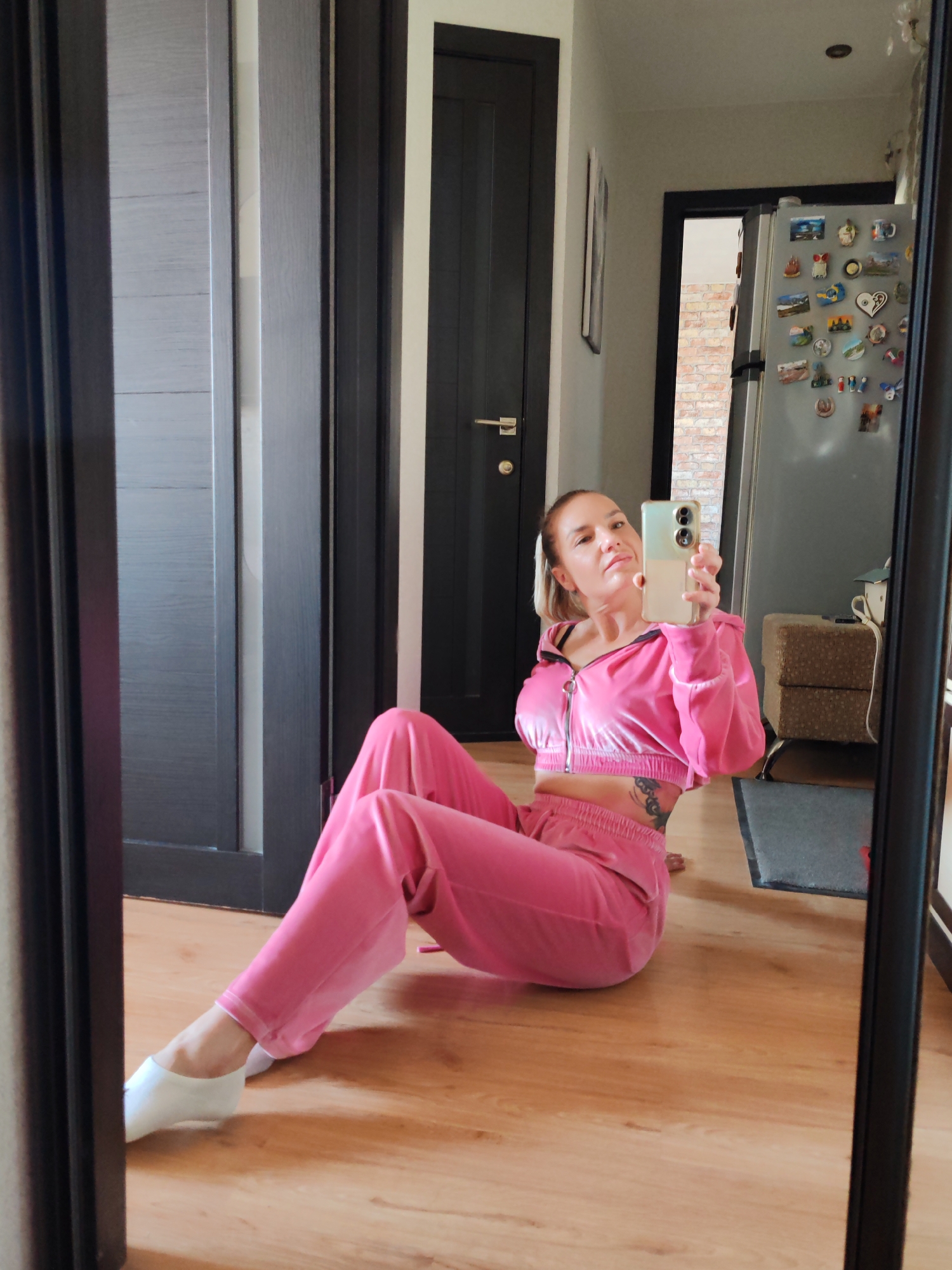 A woman is depicted in a mirror selfie, seated on a light-colored wooden floor in an indoor setting. She is wearing a matching pink velour cropped zippered hoodie and sweatpants. Her hair is pulled back, and she is holding a two-tone smartphone in her right hand, capturing her reflection. A tattoo is visible on her left side, beneath the cropped top. White slip-on footwear is visible on her left foot. The background, reflected in the mirror, shows multiple dark-colored doors, one partially open revealing a reddish-brown brick wall. A silver refrigerator covered with numerous magnets is visible to the right, reflecting overhead lighting. A stack of two textured, possibly woven, storage cubes or ottomans is positioned to the right of the woman. The room appears to have light-colored walls. The specific location of Petropavlovsk-Kamchatsky, Russia, cannot be confirmed from the visual information present in the image.