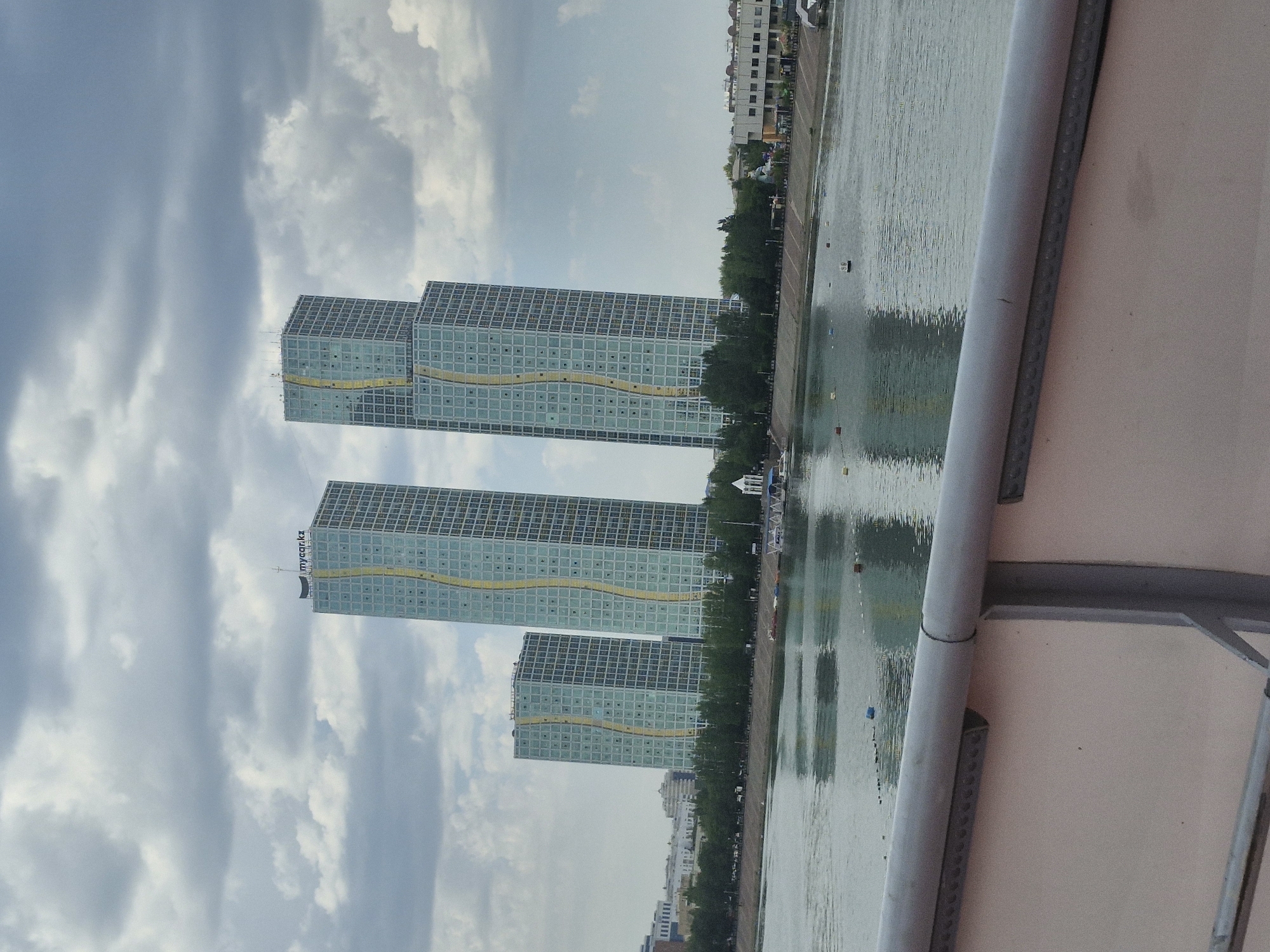The image shows a riverfront scene in Astana, Kazakhstan, with a row of modern high-rise buildings along the opposite bank. The most prominent structures are three tall, glass-fronted skyscrapers with a distinctive wavy yellow design element running vertically up their facades. One of these skyscrapers has "mycar.kz" text at its apex. The buildings appear to be residential or office complexes. In front of the buildings, there is a line of trees and a paved walkway. The river in the foreground is calm, with several small buoys and some distant boats or watercraft visible. The sky is overcast with dramatic cloud formations. A railing from a bridge or elevated walkway is visible in the foreground, obscuring the bottom portion of the scene. The overall impression is of a contemporary urban landscape with a focus on modern architecture and its integration with a natural element like the river.