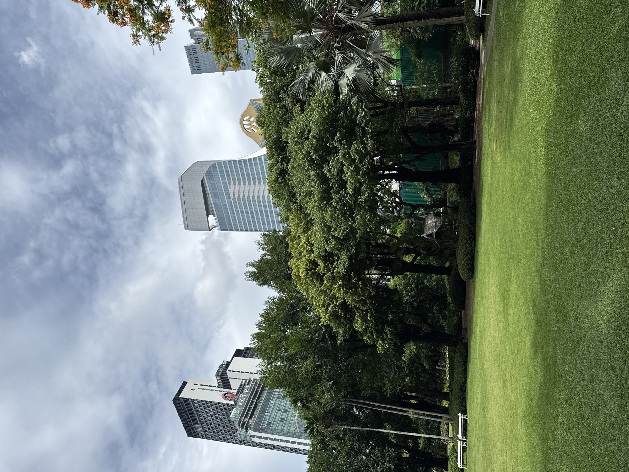 This image depicts a well-maintained park in Bangkok, Thailand, likely within the Lumphini Park area, given its blend of lush greenery and prominent skyscrapers. In the foreground, a vast expanse of vibrant green grass stretches towards the middle ground, with two white benches visible on the left side. Tall trees with dense foliage dominate the mid-ground, creating a natural barrier between the open lawn and the urban landscape beyond. Behind the trees, several modern skyscrapers rise into the partly cloudy sky. One notable skyscraper on the left features a black and white patterned facade, while others showcase contemporary architectural designs with glass exteriors. The sky is filled with a mix of white and gray clouds, suggesting an overcast day. The overall scene conveys a sense of urban tranquility, with the park providing a green oasis amidst the bustling city.