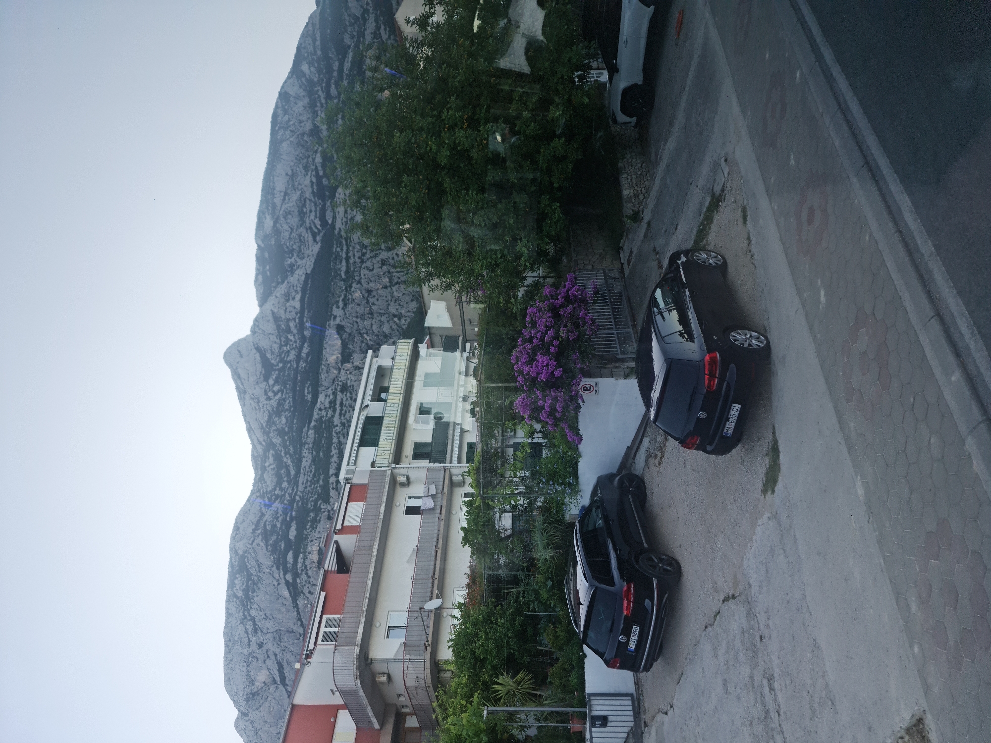 In Makarska, Croatia, a street scene unfolds with a backdrop of rugged mountains under a pale sky. In the foreground, a paved area with hexagonal stones leads to a street. Two black Volkswagen cars are parked in a gravel lot to the right. One is a hatchback, the other an SUV. Behind them, a white SUV is parked.
A multi-story building with white and red facades stands to the left, featuring balconies with railings. Lush green foliage and a vibrant cluster of purple flowers add a touch of nature. A metal fence encloses a garden area. The overall scene suggests a residential area with a mix of buildings and natural elements, characteristic of a coastal town in Croatia.