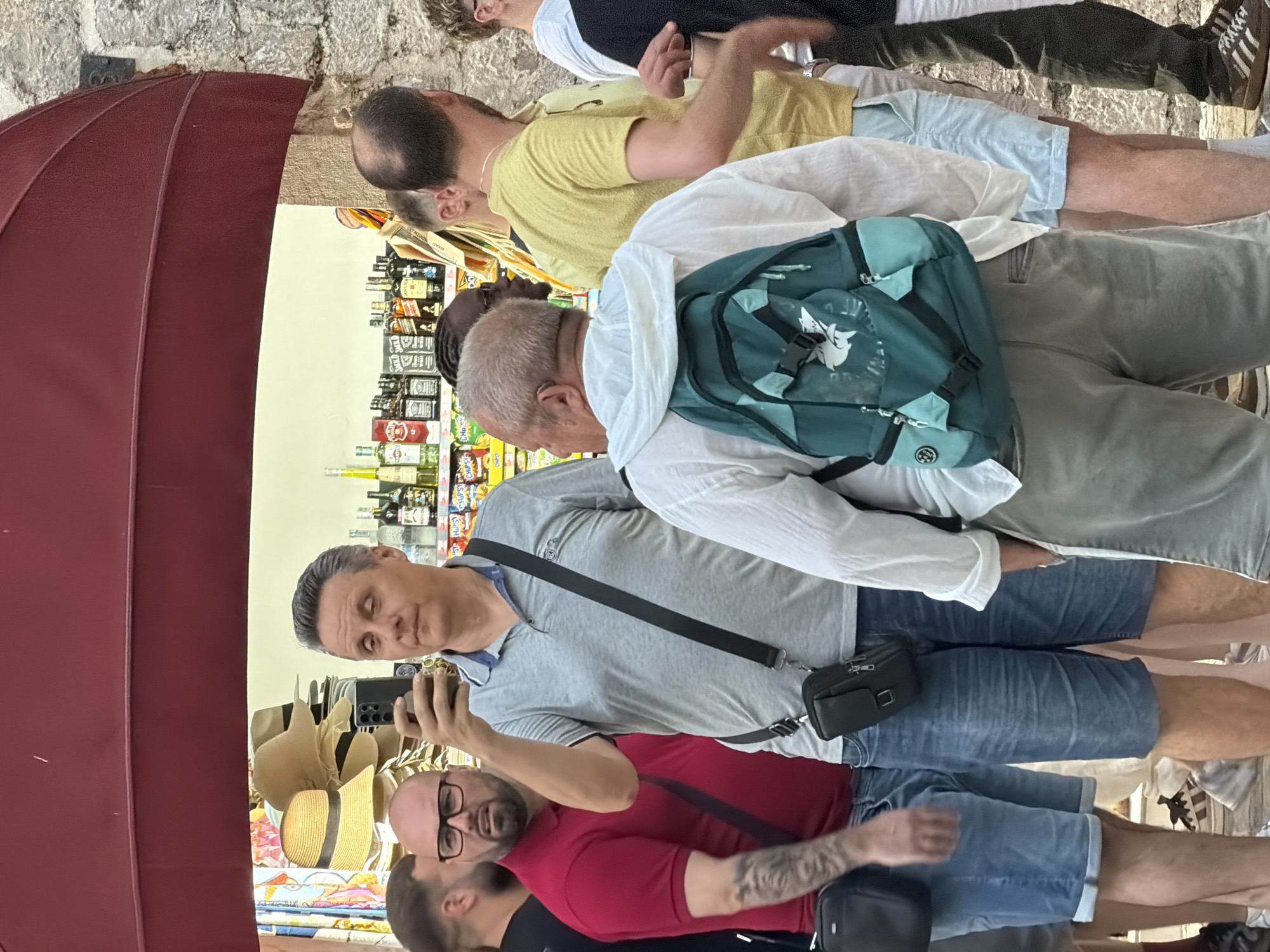 In Budva, Montenegro, a man in a grey polo shirt and denim shorts takes a selfie in front of a shop displaying straw hats and bottles of alcohol. He is wearing a black crossbody bag. To his left, a man in a red polo shirt and denim shorts looks towards the camera with a surprised expression. Behind them, other individuals are visible, including a person in a white shirt with a teal backpack and another in a yellow t-shirt. The shop appears to be an outdoor vendor or part of a marketplace, with a burgundy awning overhead and stone building materials visible to the right.