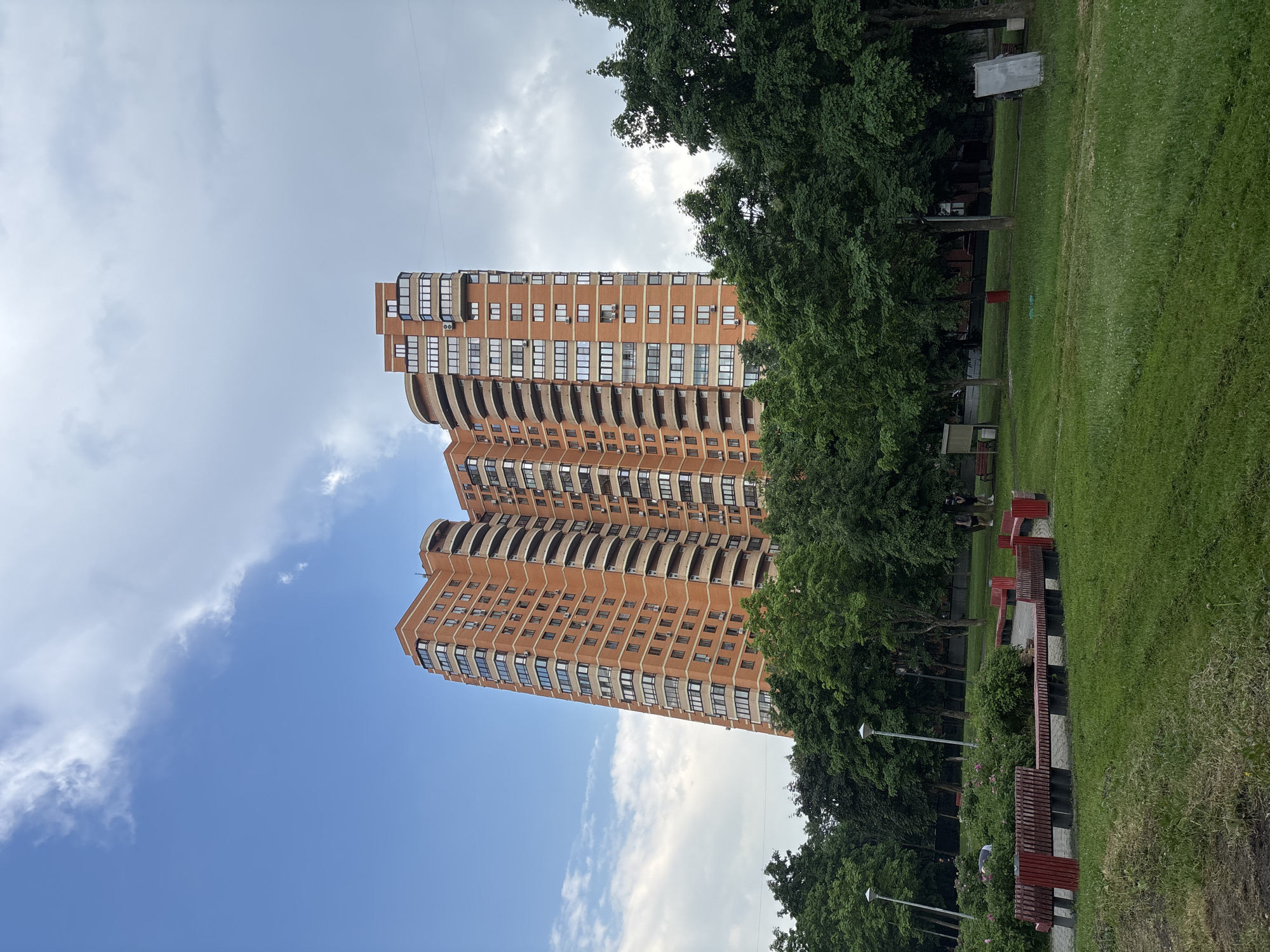 An exterior shot captures a tall, multi-story apartment building made of orange brick, situated behind a park in Moscow, Russia. The building features numerous windows and curved balconies. In the foreground, the park is comprised of a grassy lawn, a paved path, red and dark-colored benches, and lush green trees and bushes. The sky is a mix of blue with scattered white and gray clouds. Two individuals are visible in the middle ground of the park, standing near a bush.