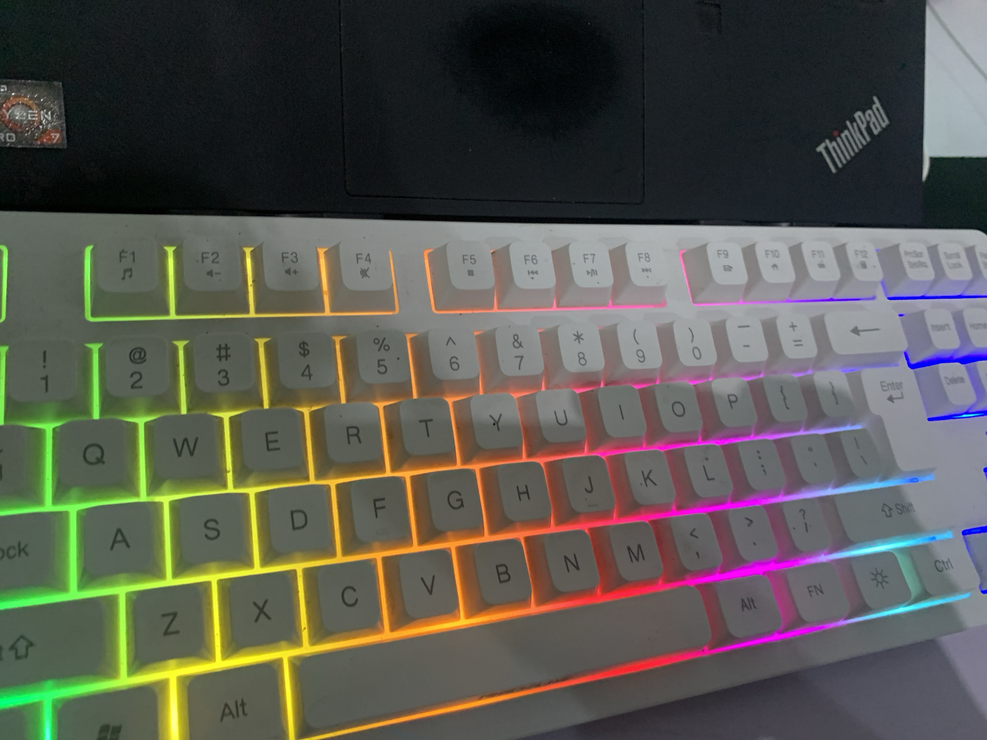 The image shows a white keyboard with RGB lighting, placed on a black surface, likely a desk. The keyboard is angled, with the top-left corner closer to the viewer. The keys are illuminated with a gradient of colors from red to green, yellow, and blue, moving from left to right. Visible keys include letters like Q, A, Z, X, C, V, B, N, M, and numbers 1 through 9. Function keys F1 through F12 are also visible on the right side of the keyboard. A logo that appears to be "AMD Ryzen" is partially visible on the black surface to the upper right of the keyboard. The ThinkPad logo is visible on the black surface to the right of the keyboard. The image does not provide any information about the location in the city of Dukuh Mojo, Indonesia.