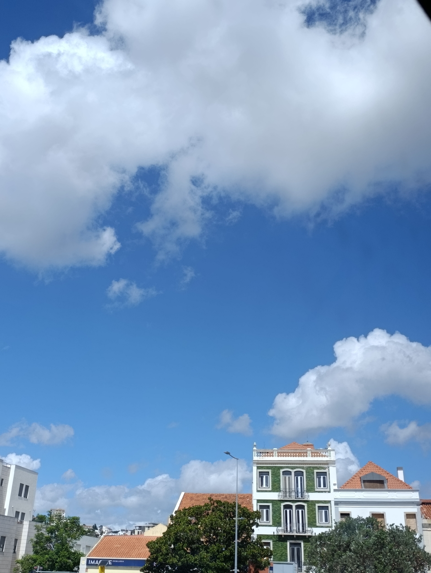 This is a ground-level, outdoor, eye-level shot of a street scene in Setúbal, Portugal. The bright blue sky is filled with fluffy white clouds, with a large cloud formation dominating the upper portion of the frame. In the lower half of the image, several buildings are visible. On the left, a modern white building with dark windows stands partially in frame. To its right, a distinct building with a dark green tiled facade and white trim is prominent. This green building features multiple windows, balconies with dark railings, and a white decorative cornice along the top, topped with a terracotta tiled roof. Adjacent to the green building on the right, another building with a terracotta tiled roof and white facade is partially visible. In the foreground, the tops of lush green trees obscure the lower parts of the buildings. A lamppost is positioned to the left of the green building. A yellow sign with black lettering, partially obscured by trees, reads "IMÁGINE IMOBILIÁRIA." The overall scene suggests a sunny day in a city.