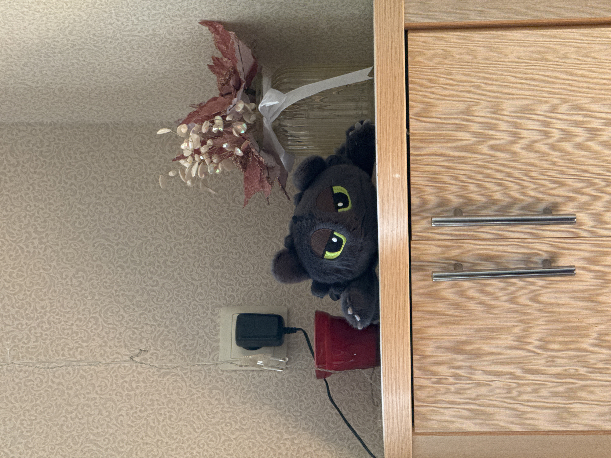 The image shows a stuffed toy of Toothless from How to Train Your Dragon lying on a wooden shelf. To the left of the toy is a red pot with a black wire extending from it, plugged into a black power adapter that is connected to a wall outlet. Above the shelf, the wall is covered with textured wallpaper. To the right of the toy, there is a glass vase filled with decorative flowers and a white ribbon tied around its neck. The shelf is above two wooden cabinet doors, each with a silver handle. The location is not specified in the image.