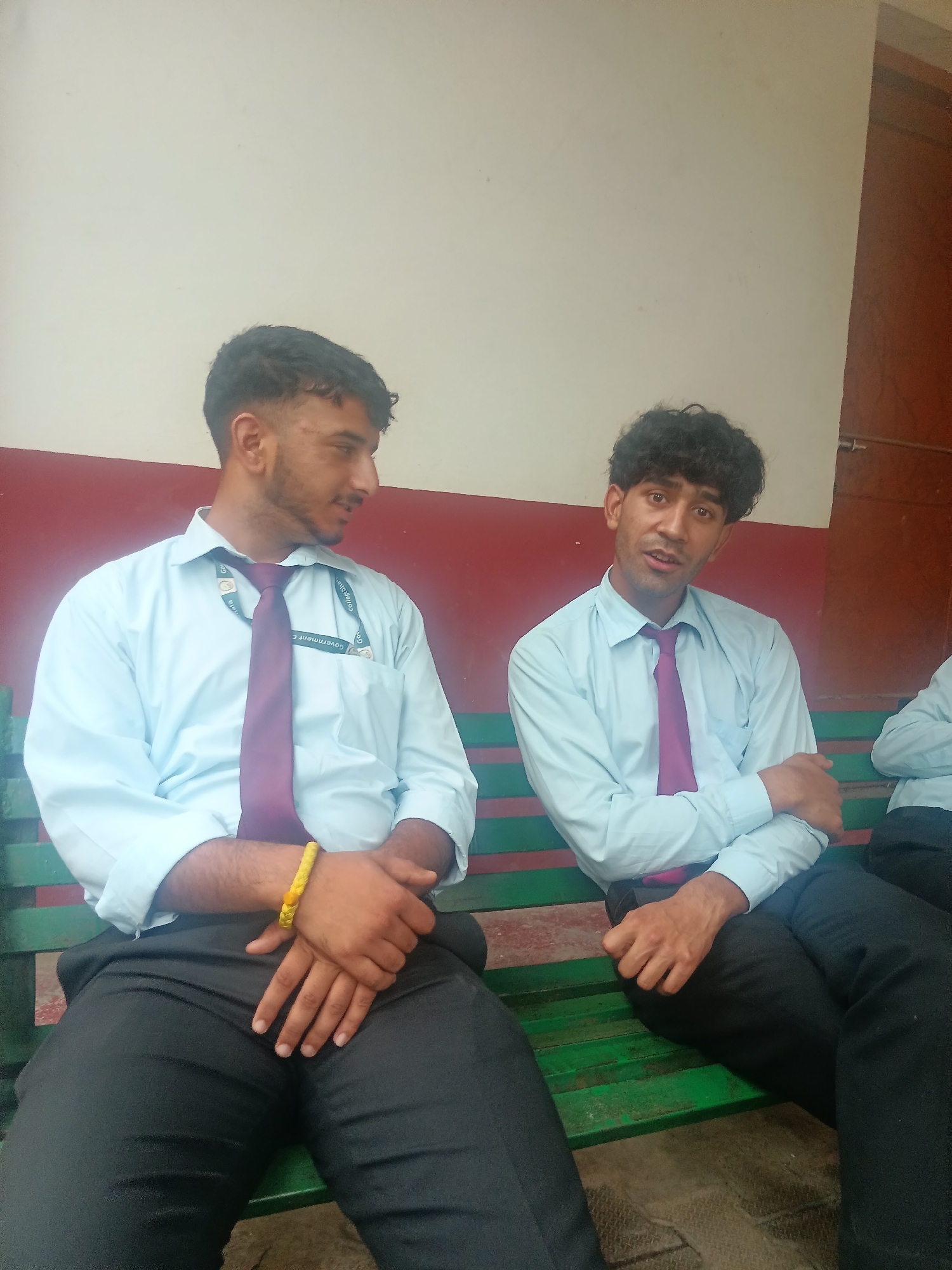 Two young men, possibly students, are seated side-by-side on a green wooden bench against a red and cream-colored wall. Both are wearing light blue collared shirts with dark ties and black pants. The man on the left is looking to his right, with his arms crossed over his lap. He has a yellow bracelet on his left wrist. The man on the right is looking directly at the camera and speaking, with his left arm bent and resting on his knee, and his right arm slightly extended. A portion of a third person is visible on the far right of the image, also seated on the bench. The overall setting suggests an outdoor or semi-outdoor environment in Dharamshala, India, given the architecture and the presence of what appear to be identity cards or lanyards around their necks.