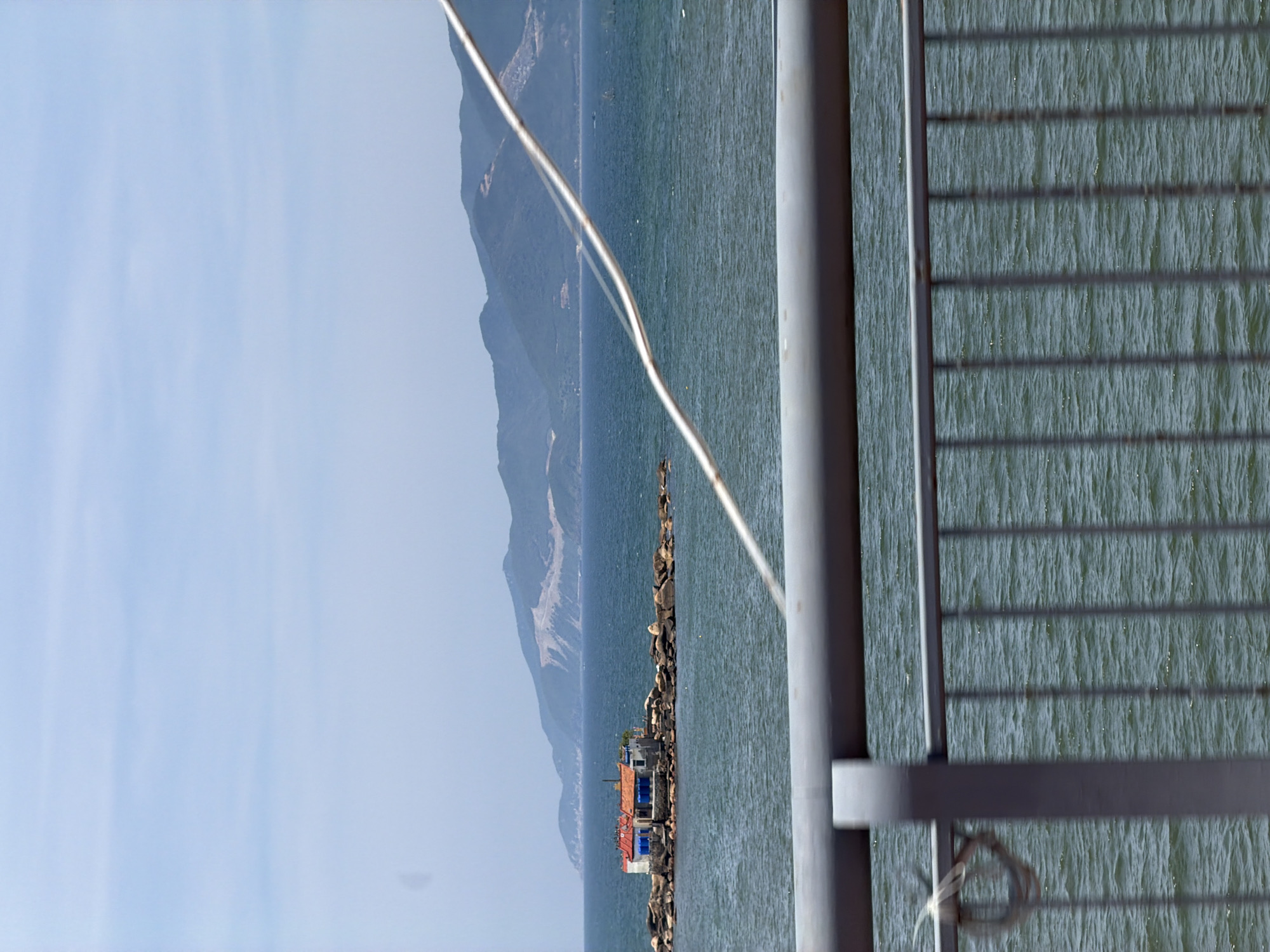 A small, multi-colored building with red roofs and blue windows sits on a rocky outcrop in the ocean. The building is surrounded by dark blue water, with a range of green, hazy mountains in the background under a pale blue sky. In the foreground, a grey metal railing runs horizontally across the bottom of the image, with vertical bars extending down, partially obscuring the view of the water. A white wire is visible diagonally across the scene, connecting from the upper right to the mid-left. The scene appears to be captured from a moving vehicle, possibly a bridge or a ferry, given the blur of the railing and the angle of the shot. The location is identified as Bac Nha Trang Ward, Vietnam.