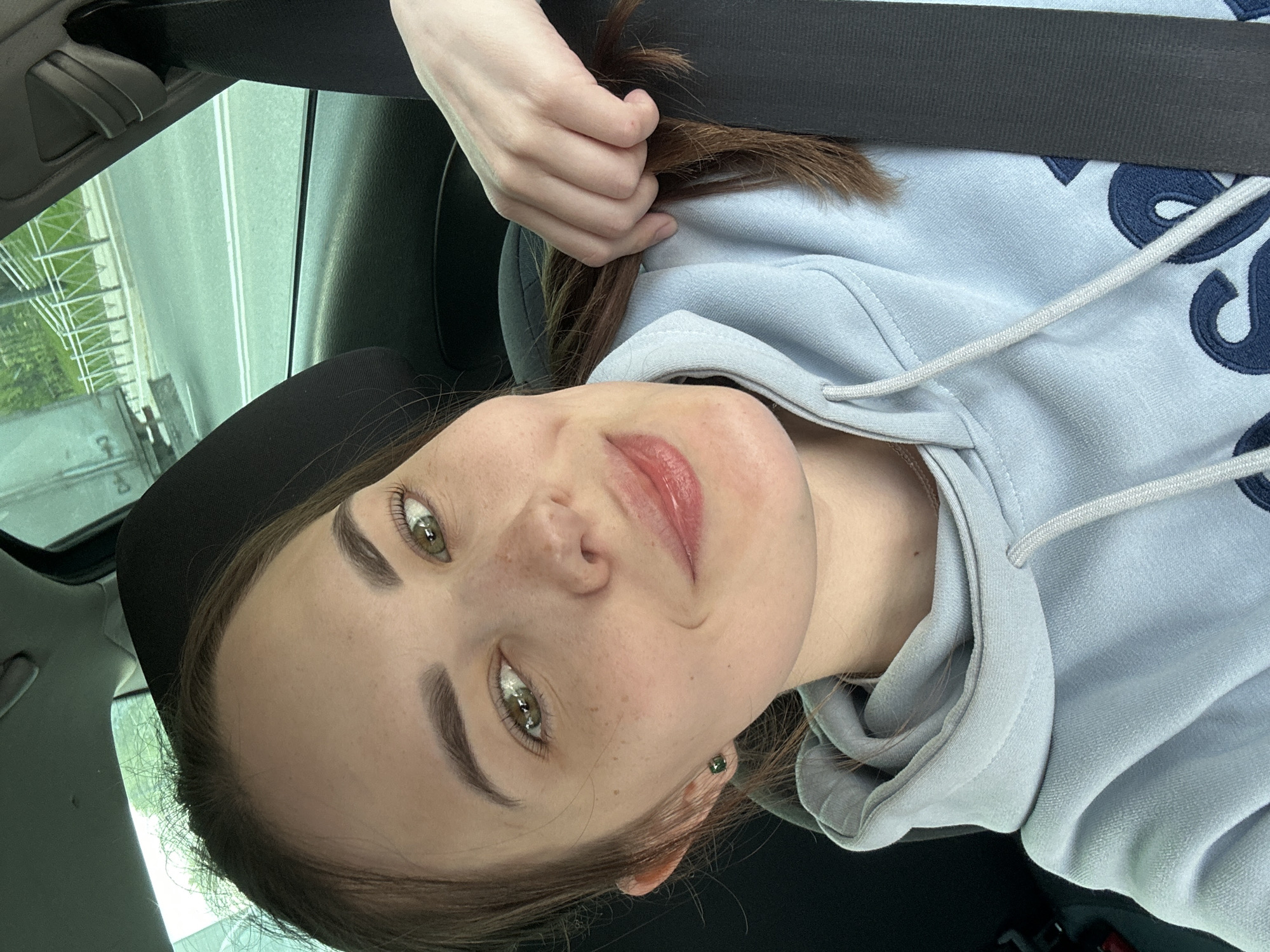 A selfie taken in a car shows a young woman wearing a light blue hoodie with blue lettering and a seatbelt. She is holding a strand of her brown hair. Through the car window, a road and some green vegetation are visible. The photograph was taken in Perm, Russia.