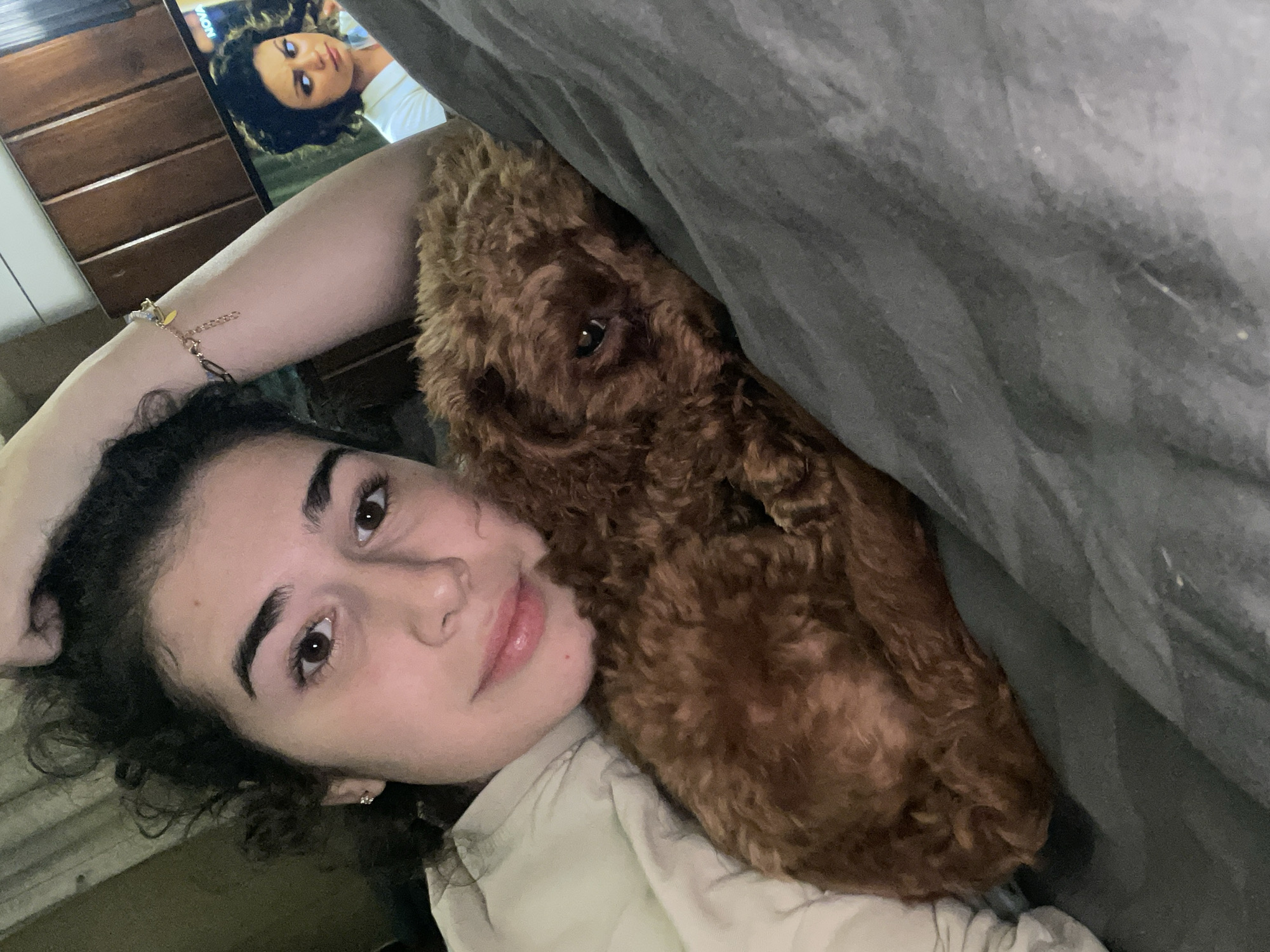 A young woman is lying down next to a brown, curly-haired dog, possibly a poodle, under a gray blanket. The woman is looking directly at the camera. Her face is visible, and she has dark hair and eyebrows. She is wearing a light-colored shirt or jacket. The dog is positioned close to her head, and its eye is visible. Behind her, a television screen shows a face, possibly from a movie or show. To the left of the screen, a wooden dresser or cabinet is visible. No indication of Sofia, Bulgaria is visible.