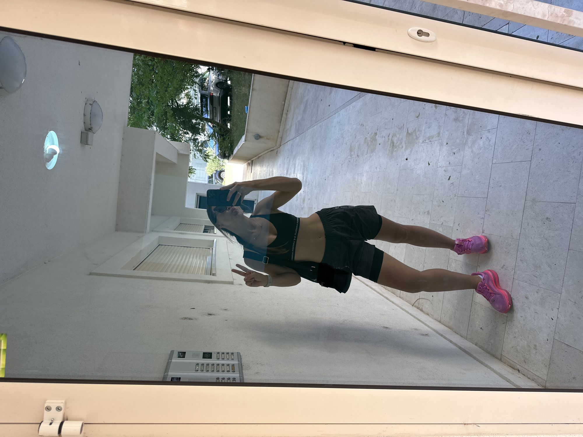 A woman, reflected in a full-length mirror, stands in a paved outdoor walkway. She is wearing a black sports bra, black shorts, and pink athletic shoes. She holds a black bag over her left shoulder and is using her right hand to take a selfie with her phone, while her left hand is raised in a peace sign. The background visible in the mirror includes a building with closed shutters, overhead lighting fixtures, trees, and parked cars. The mirror is framed by white and beige columns. The setting appears to be an apartment complex or residential area in Boreti, Montenegro.