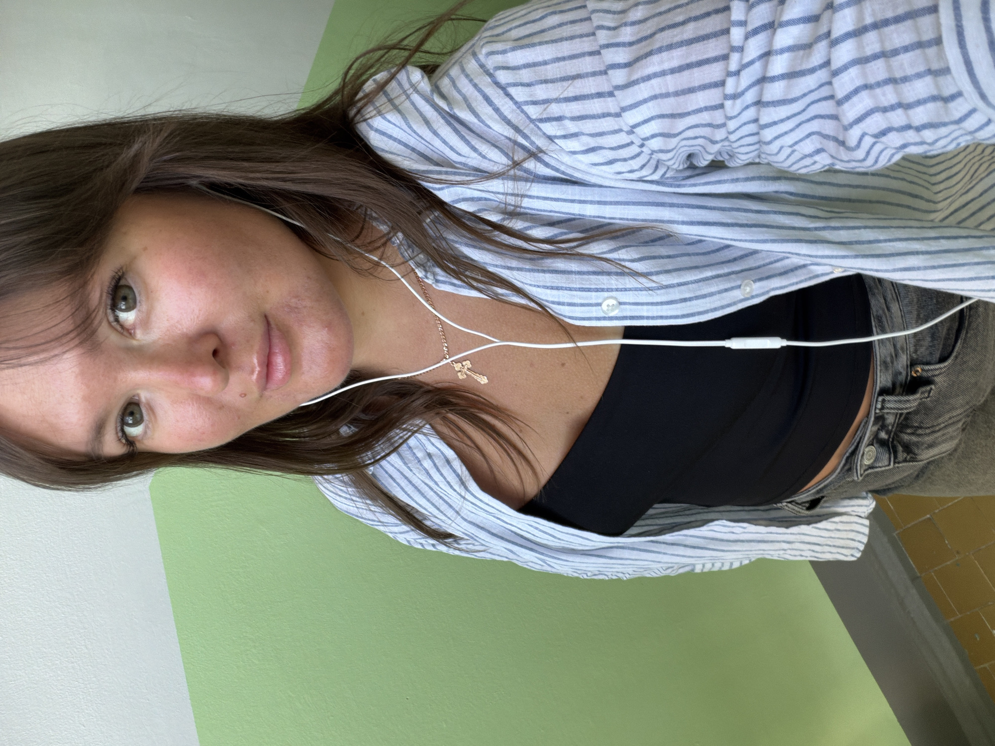 A woman is standing indoors against a wall that is painted half white and half light green. She has brown hair and is wearing a light blue and white striped shirt over a black top, and gray denim shorts. She also wears a gold necklace with a cross pendant, and white earphones are visible. Part of a tiled floor is visible on the right edge of the frame. The location in the city of Chaikovsky, Russia, cannot be determined from the provided image.