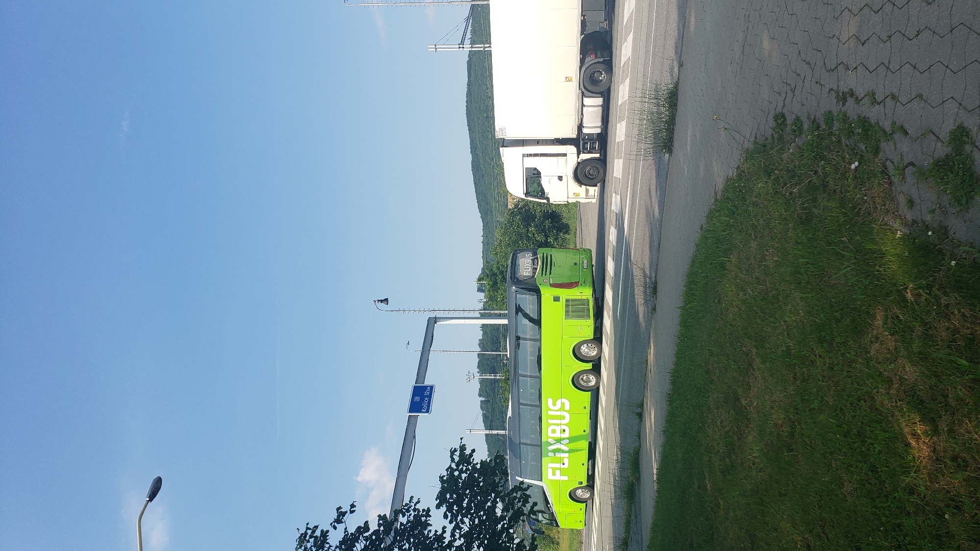 In Prešov, Slovakia, a bright green FlixBus coach, positioned on a pedestrian crossing, is intersected by a white truck on a sunny day. The FlixBus, featuring a prominent white "FLIXBUS" logo, has its rear facing the camera, with its front moving to the left. To its right, the white truck, with its trailer attached, is moving in the same direction. The road is marked with white lines and has a paved sidewalk to the right, bordered by a grassy area. Overhead, a blue road sign indicates "Košice 32km". A streetlamp is visible on a pole to the right of the FlixBus. The background shows trees and rolling hills under a clear blue sky.