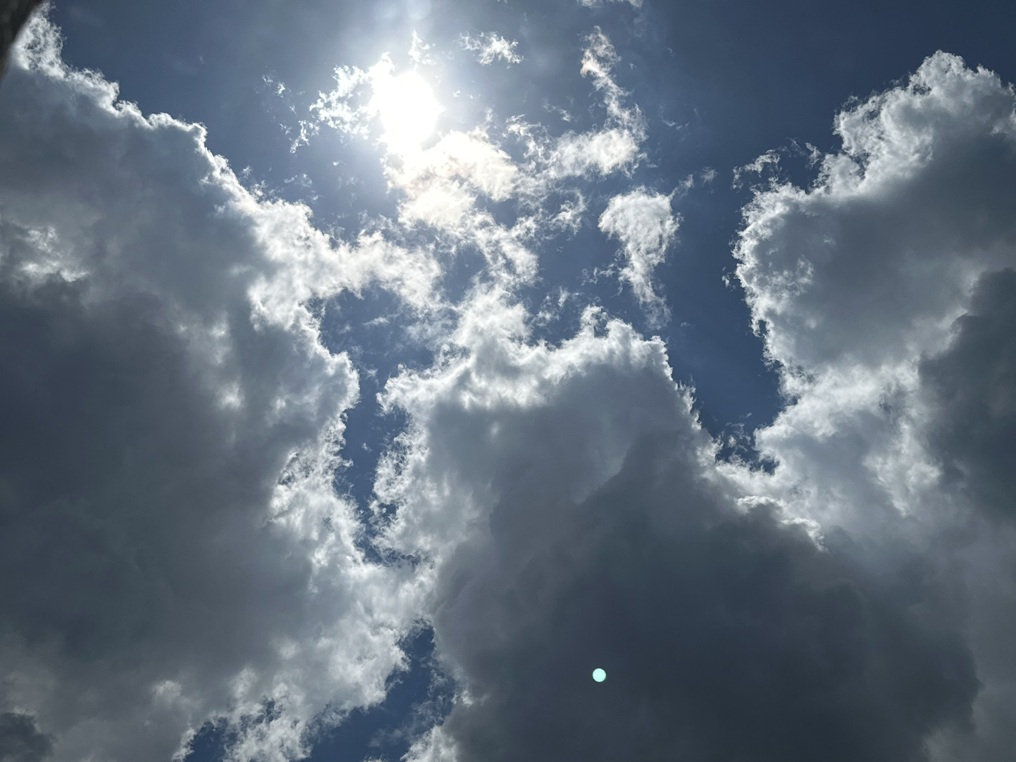This is a low-angle, close-up, full-shot photograph of a cloudy sky. The sun is a bright, blown-out white orb near the top of the frame, partially obscured by wispy white clouds that are illuminated by its rays. Below these bright clouds are larger, darker, and more voluminous grey clouds that occupy most of the lower half of the image. The sky is a deep blue, visible through gaps in the clouds. In the lower right quadrant of the image, a small, cyan-colored bokeh-like circle is visible. The scene is devoid of any human presence or man-made structures.