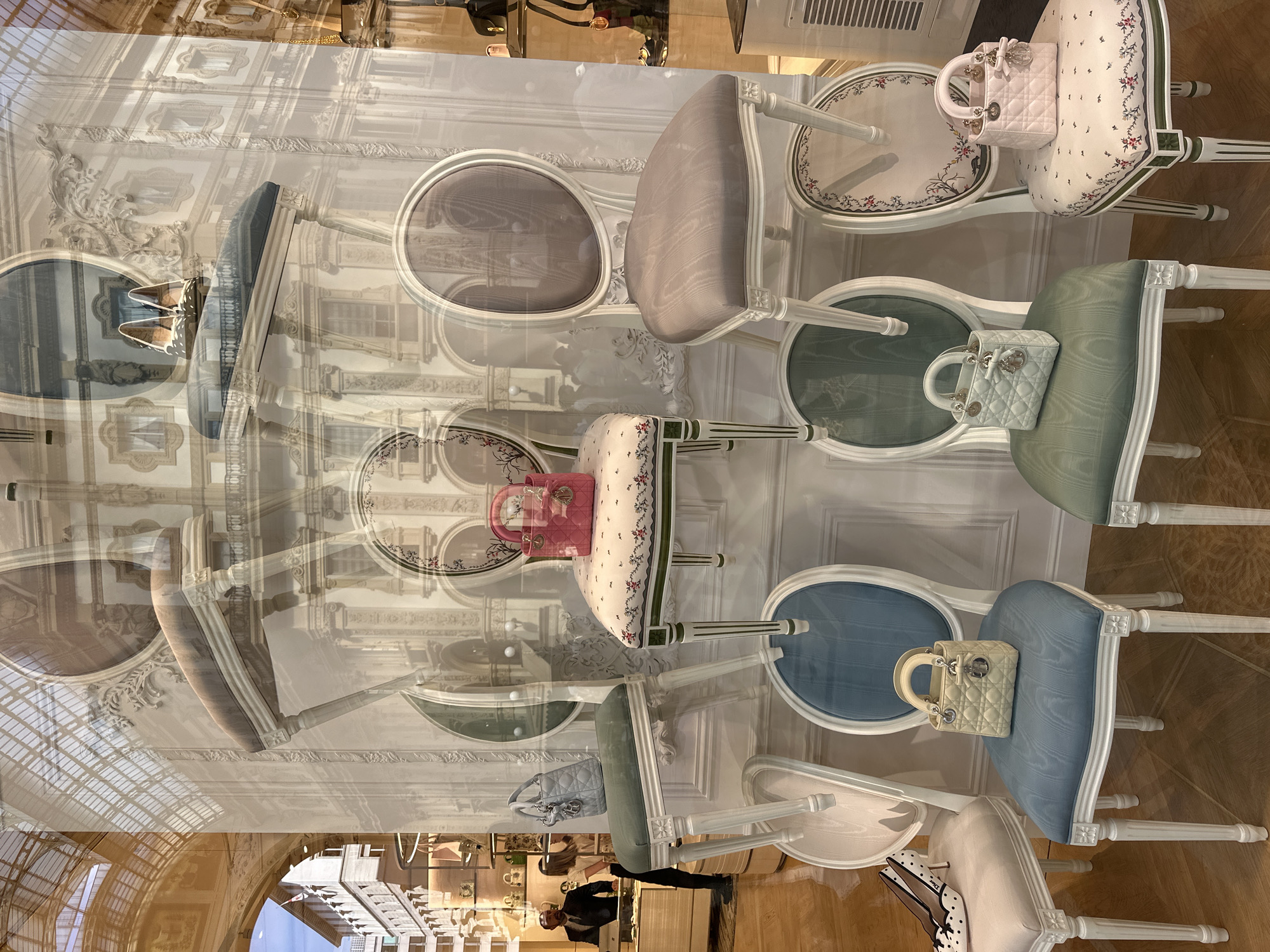 The image depicts a luxury boutique window display in Milan, Italy. The display features several ornate chairs with upholstered seats in various colors including white, light blue, and green. Each chair is accessorized with a small, quilted designer handbag, primarily in pastel shades like pink, blue, and beige. One chair is also adorned with a pair of black and white polka dot high-heeled shoes.
The background of the display is a reflection of the surrounding Galleria Vittorio Emanuele II, a historic covered arcade in Milan known for its high-end shops and cafes. The reflections show architectural details of the Galleria, including vaulted glass ceilings, ornate facades, and balconies. In the lower left corner of the image, a man in a suit and a woman are visible through the display window, suggesting they are customers or staff inside the store. The lighting is bright, typical of a retail environment, and the overall scene conveys an atmosphere of luxury and elegance.