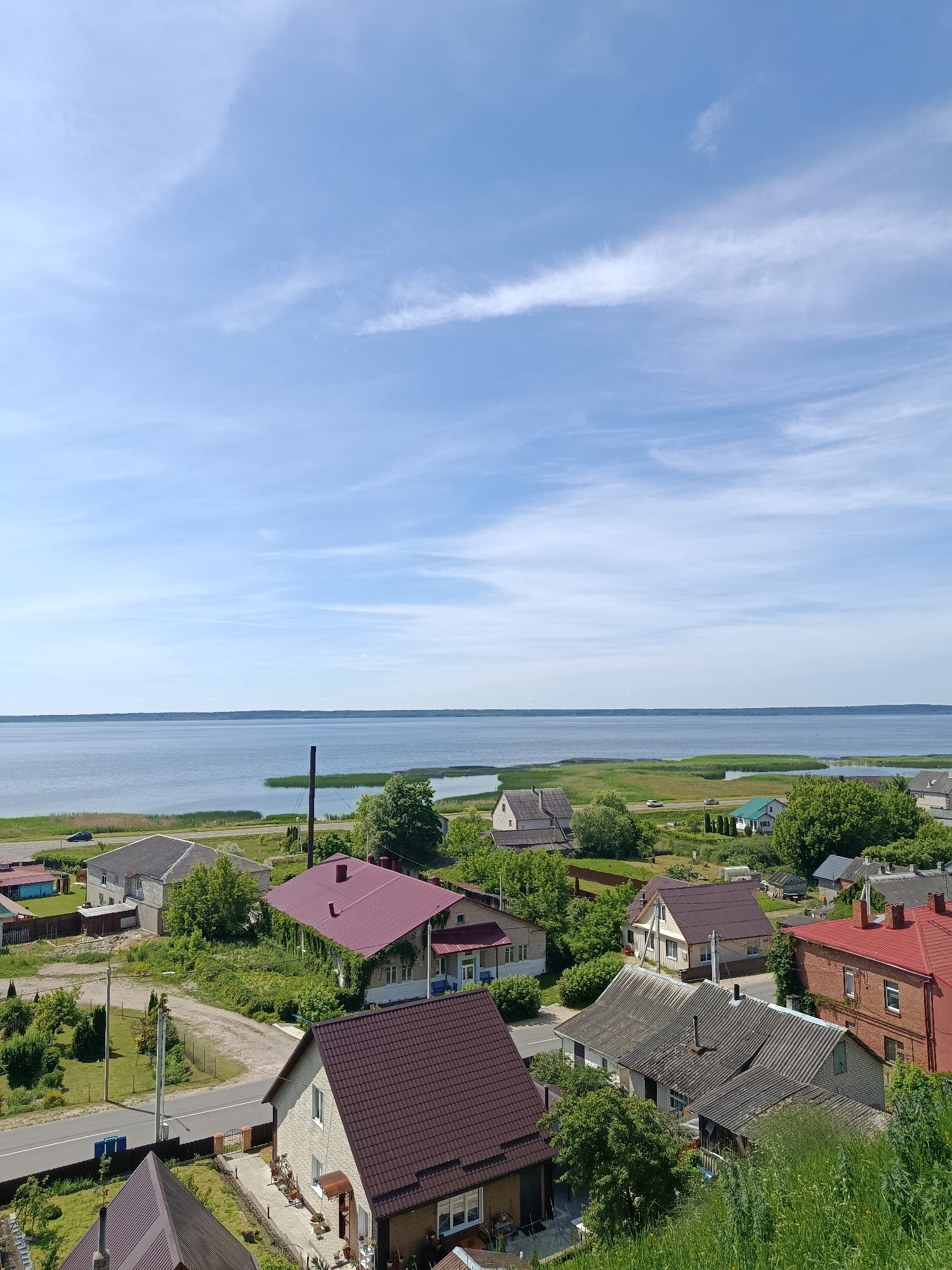 The image captures a panoramic view of a lakeside settlement, likely in Braslaw, Belarus, on a clear, sunny day. The foreground is dominated by the roofs of several houses, varying in color and architectural style, nestled amidst lush greenery. One prominent house features a striking deep red tiled roof, with ivy climbing its exterior walls. Another house, closer to the viewer, has a beige textured facade and a dark brown tiled roof, with a paved walkway leading to its entrance, adorned with potted plants and outdoor furniture.
Beyond the houses, a road meanders through the settlement, with a few cars visible. The mid-ground is characterized by a strip of vibrant green reeds and trees lining the edge of a vast, calm body of water, presumably Lake Drivyaty, given the location context. The lake stretches out to the horizon, meeting a pale blue sky with wispy white clouds. A solitary dark chimney-like structure rises from the reeds near the water's edge. The overall scene depicts a tranquil rural or semi-rural landscape, typical of a lakeside town or village.