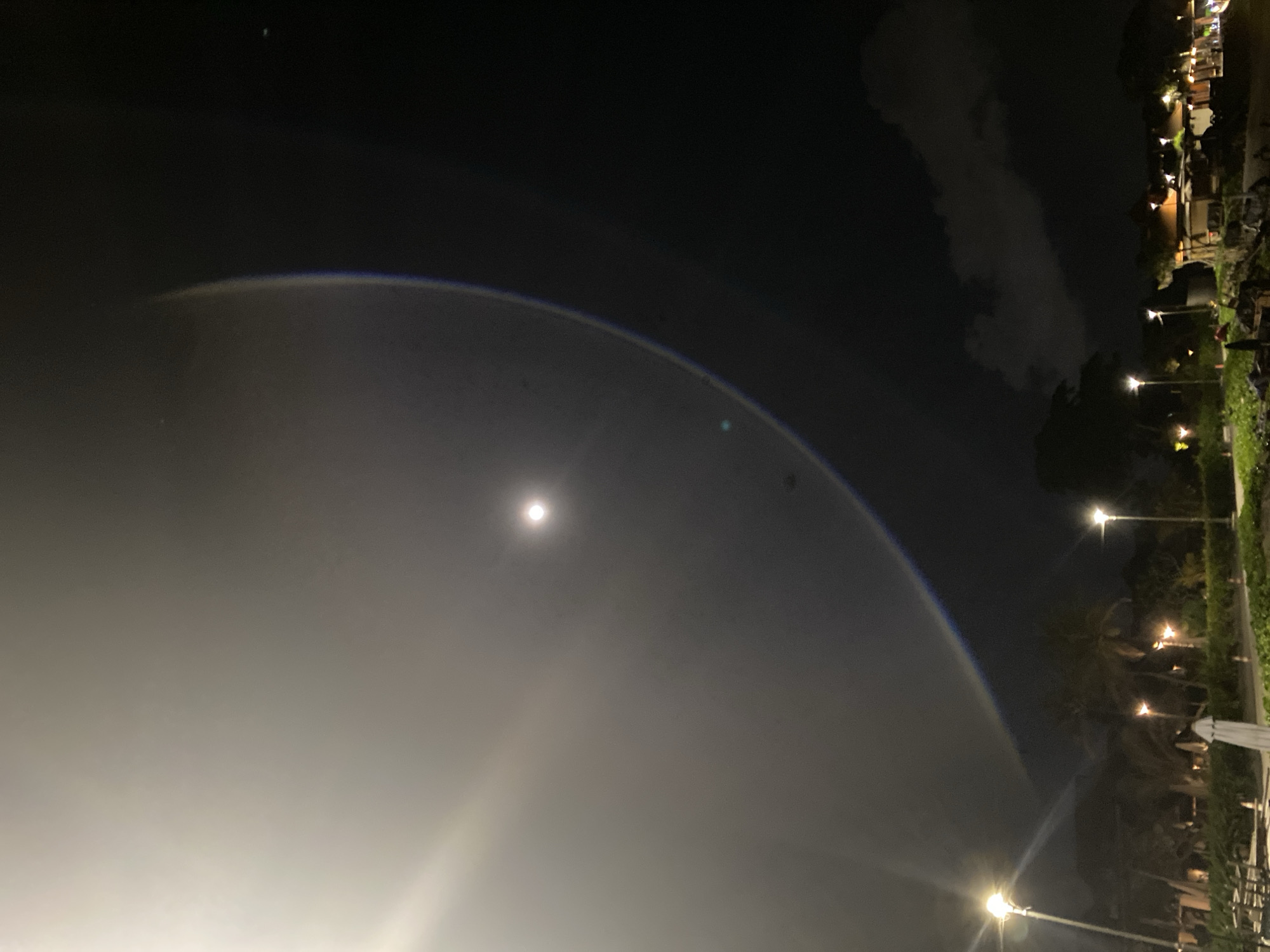Captured at night in North Kuta, Indonesia, the image presents a scene of a lunar halo. The moon appears as a bright orb in the center of the halo, which manifests as a faint, broad arc across the sky. To the right of the frame are various lights indicating buildings and streetlights, scattered amid the vegetation. The night sky is dark, with some clouds visible. The perspective is a ground-level shot. The picture shows a nighttime scene with minimal human presence, emphasizing the atmospheric phenomenon and urban lighting.