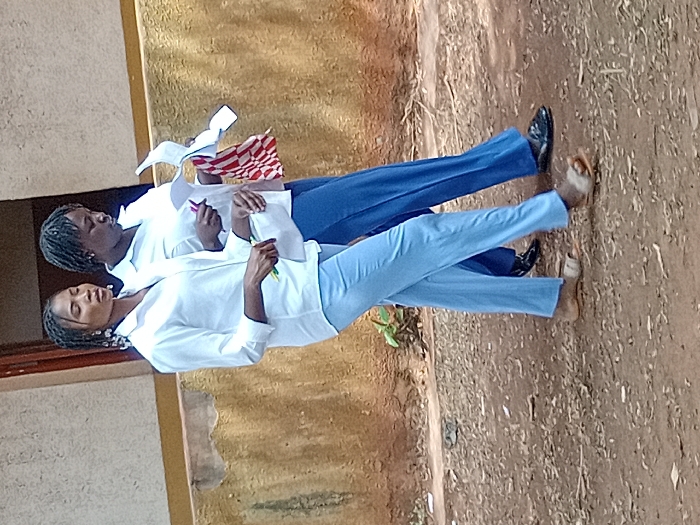 Two students stand outdoors in front of a building with a faded yellow facade. Both wear white, long-sleeved shirts. One has on blue pants and polished shoes, and is holding papers and a checkered cloth. The other student is wearing light blue pants and sandals, with a writing utensil in hand. The ground is bare earth with some small plants.