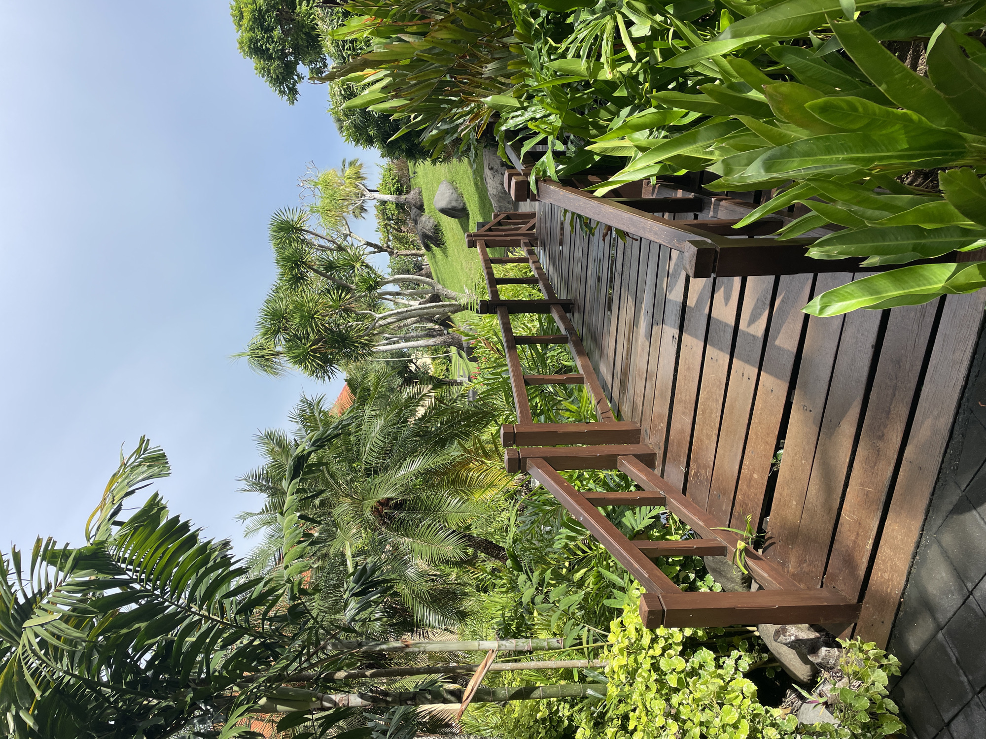 The image depicts a wooden bridge or walkway with brown handrails in a lush, tropical garden setting. The bridge is constructed of parallel wooden planks, and the handrails feature a ladder-like design. The surrounding environment is dense with various types of tropical plants, including palm trees, broad-leafed plants, and other greenery. A clear blue sky is visible above the trees. The photograph was taken in Kuta Selatan, Indonesia, based on the surroundings that correspond to the flora and characteristics of the city. The scene portrays the natural and possibly manicured landscape of this area.