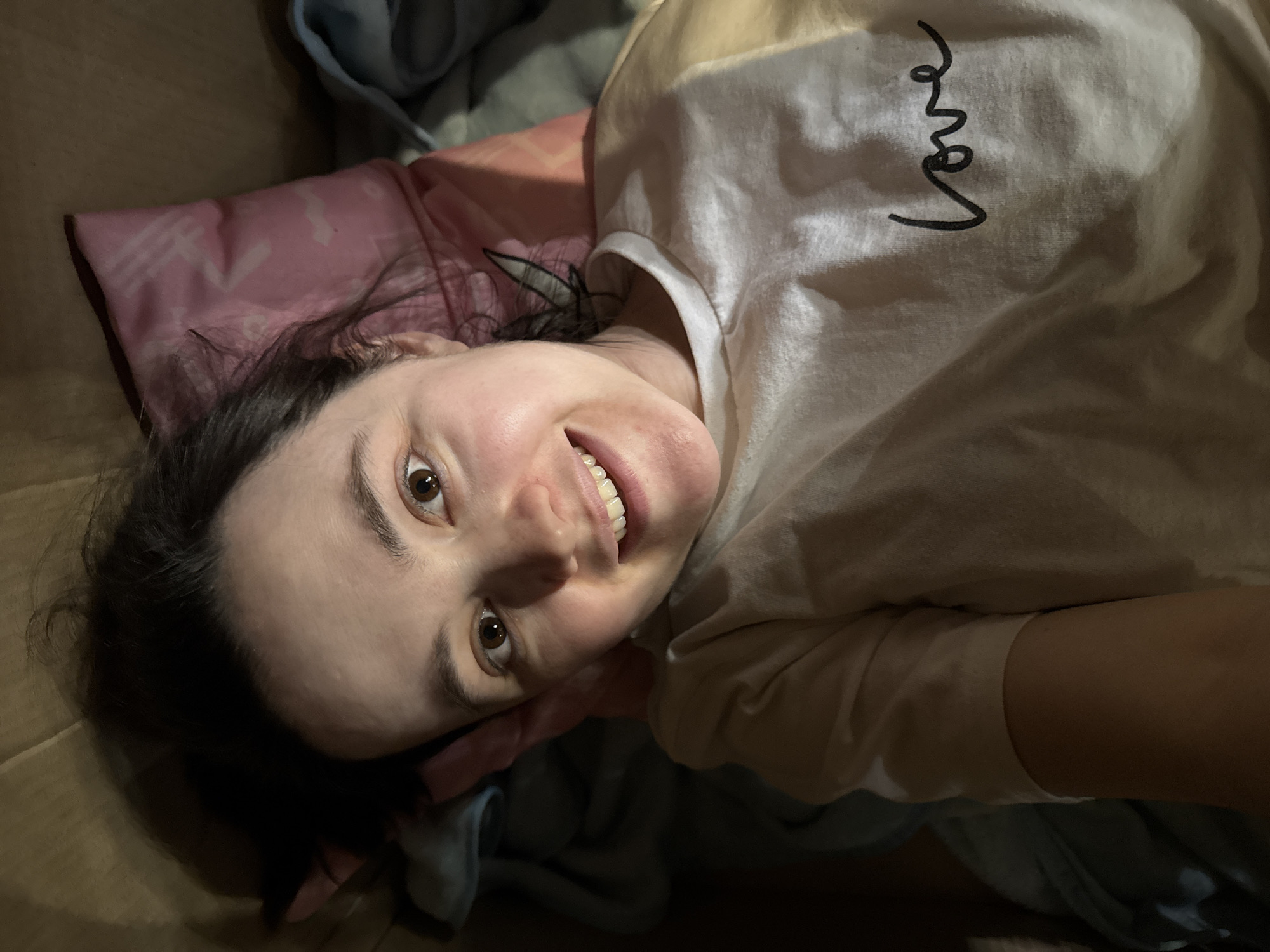 A woman is lying down and smiling at the camera. She is wearing a white t-shirt with the word "love" written on the shoulder. She is surrounded by pillows and blankets. It is not possible to ascertain the specific location in Izhevsk, Russia from the image.