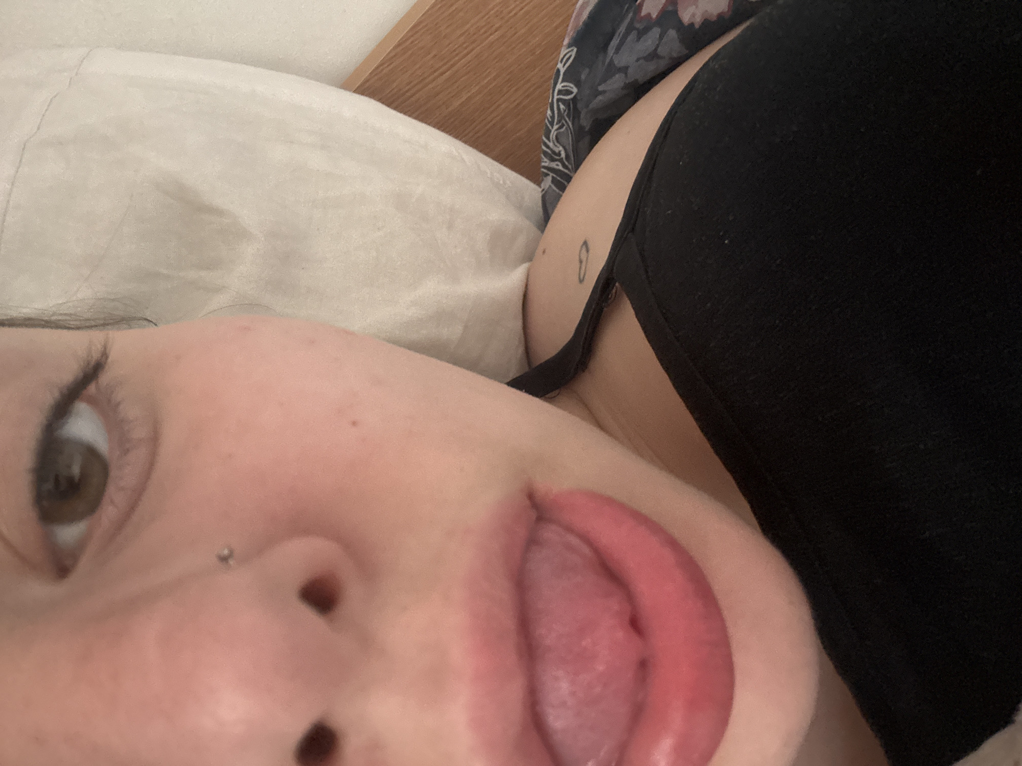 A close-up, slightly tilted, selfie-style image depicts a person lying on a white pillow, possibly in bed. The person has fair skin, brown hair, and hazel eyes with a piercing on their left nostril. Their tongue is partially extended. They are wearing a black top, and a small heart tattoo is visible on their upper chest or shoulder. The background includes a wooden headboard and floral patterned bedding. The location is unknown and cannot be determined to be Troyan, Bulgaria, from the information provided.