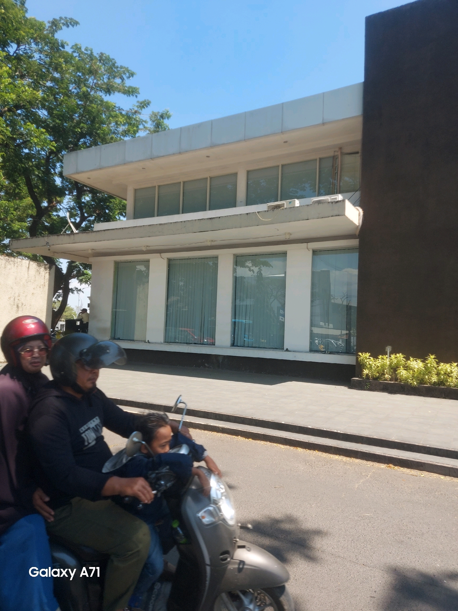 The image captures a street scene in Tombolo, Indonesia, under a clear blue sky. In the foreground, a scooter with three occupants is moving from left to right. The driver, wearing a black helmet, is partially obscured. Behind him, a person wearing a red helmet and glasses is seated, and a child is positioned between them. The scooter is dark gray with silver accents.
To the right of the scooter is a paved sidewalk and a road. Beyond the sidewalk, a modern, two-story building with a dark, imposing facade on the right and a white section with large windows on the left is visible. The white section features several large glass windows with vertical blinds, and an overhang with air conditioning units. A lush green tree with dense foliage is situated to the left of the building. The overall impression is a bright, sunny day in an urban or semi-urban environment.