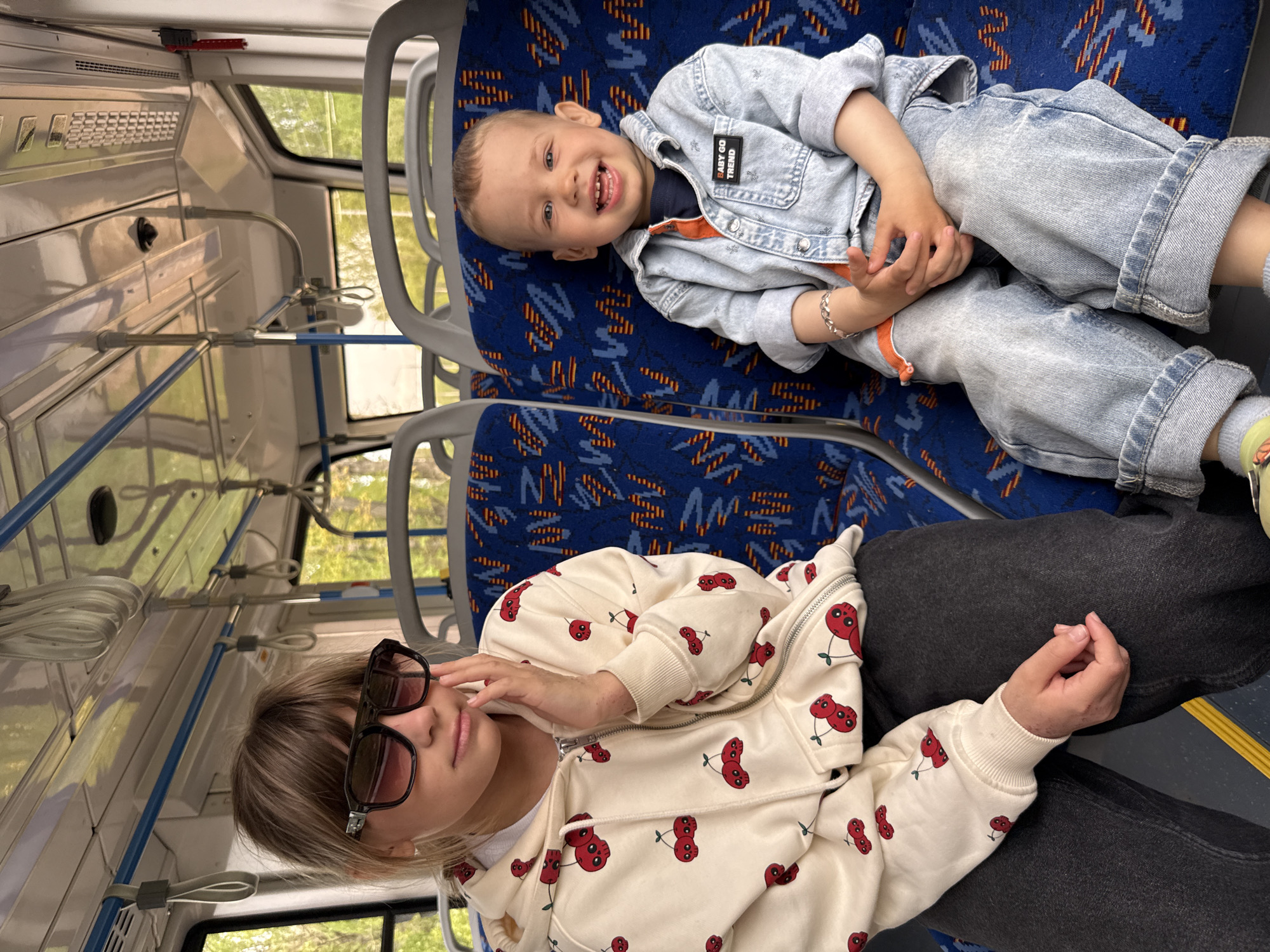 A photograph taken inside a vehicle depicts two individuals. The younger individual, presumably a child, is lying on the upper seat. This child is wearing a denim jacket with a patch that reads "BABY GO TREND" and is smiling. Adjacent to this child, on the lower seat, is an individual wearing a light-colored hoodie with a cherry pattern. This person is wearing sunglasses and is holding their hand near their face. The seats are patterned with a design primarily in blue. The environment appears to be a bus or similar public transportation vehicle, with handrails and windows visible. The location is Unknown, Unknown.