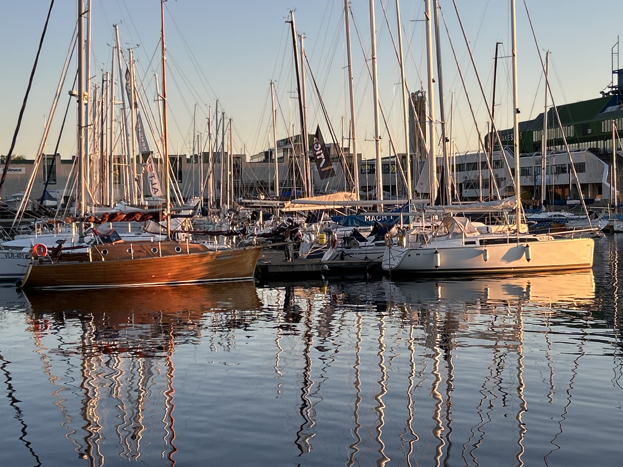 This image depicts the Tallinn Old City Harbour (Vanasadama Jahisadam) in Estonia during the golden hour, characterized by warm, low-angle sunlight. The scene is dominated by numerous sailboats docked in the marina, their tall masts and intricate rigging forming a dense vertical pattern against the sky.
In the foreground, to the left, a classic wooden sailboat with a rich, reddish-brown hull is prominently featured, reflecting the warm light. A red life buoy is attached to its stern. Faint lettering, possibly "SIRIUS," is visible on its stern. To the right in the foreground, a modern white sailboat with a sleek hull also catches the golden light. The registration number "VLR-401" is visible on the hull of a white boat positioned behind the wooden vessel. Between these boats, a wooden pier is visible, with at least one person partially obscured, standing on it.
The water surface is relatively calm, exhibiting gentle ripples that create elongated, wavy reflections of the masts, rigging, and boat hulls. These reflections are a mix of golden light, deep blue water, and shadowed browns and whites.
The midground is filled with many more sailboats, their masts rising uniformly. A black banner displaying the letters "UICA" (appearing reversed, "ACIƱ," due to its material) hangs from one of the masts. A blue canvas cover on a nearby boat features the text "MACH I".
The background consists of several buildings. On the left, partially obscured by masts, a building facade displays the partial word "Sailinvest." Further left, a tall, slender, dark structure, likely a tower or part of a larger complex such as Linnahall, rises above the other buildings. In the center, light-colored industrial or office buildings with multiple windows are visible. Text, including "VANAKES S" and "GUN LA TIR," appears on some of these facades or associated signs. To the right, a large, elongated building with a distinctive green roof and large windows, possibly a port terminal or warehouse, completes the urban backdrop.
The sky above is a clear gradient, transitioning from a light blue at the zenith to a warm, golden hue near the horizon, indicating either sunrise or sunset. No active sailing or significant human interaction is visible; the boats are static, moored at the docks.