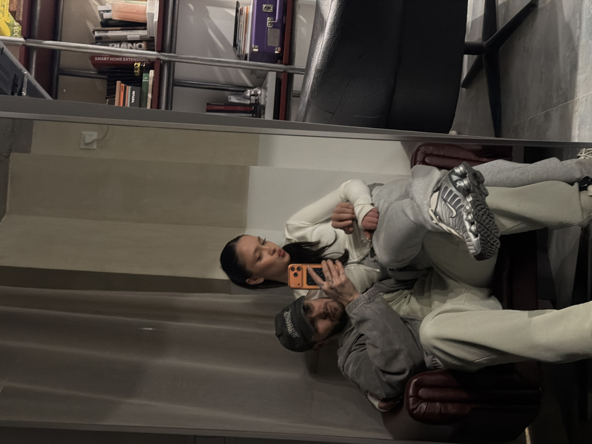 A reflection of a couple taking a selfie is visible in a mirror. The woman is wearing a white top and the man is wearing a gray hooded jacket and a baseball cap. They are holding an orange smartphone. They are sitting on a red chair. The background includes shelves filled with books and objects, and a black chair. The location is unknown, and there is no information to suggest that it is in Yerevan, Armenia.