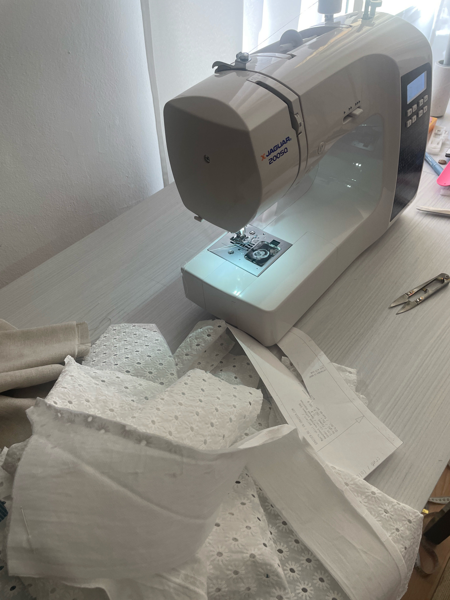 The image shows a sewing machine on a light-colored wooden desk, in Atarfe, Spain. The sewing machine is white and beige, with the brand name "X JAGUAR 20050" visible. Various pieces of white fabric, some with eyelet embroidery, are scattered on the desk in front of the machine. There are also some sewing pattern papers, a pair of small scissors, and a measuring tape. The scene appears to be a home sewing area, with bright light filtering in from a window, suggesting it is daytime. The overall impression is one of a crafting space, possibly in a residential setting. There are no people visible in the image.