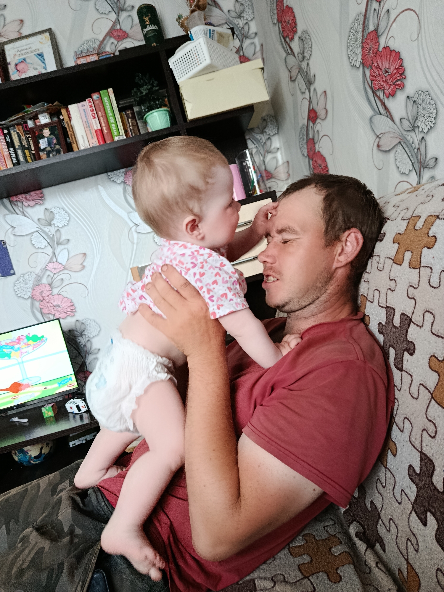 A medium shot shows a man holding a baby, presumably in an apartment. The man, wearing a red t-shirt, is reclined against a couch with a puzzle-piece pattern. He is looking at the baby who is touching his forehead. The baby is wearing a diaper and a short-sleeved shirt with a heart pattern. A desk with a computer monitor is visible to the left; the monitor displays a cartoon-like image of a martini glass. Behind the man and baby are a bookshelf filled with books and decorations and wallpaper with a floral pattern. Based on the location data, this is in the city of Фоки, Russia.