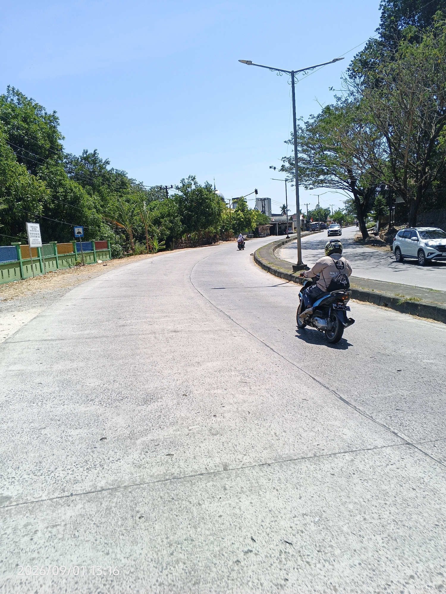 The image depicts a sunny day on a paved road in Kupa, Indonesia, likely a suburban or semi-urban area. The road curves to the right, with a concrete embankment on the left and a slightly elevated asphalt surface on the right. Lush green trees line both sides of the road, with some appearing to be tropical species. A double-headed street lamp stands prominently on the right side, casting a shadow on the road.
Several vehicles are present. In the foreground, a person wearing a light-colored shirt, dark pants, and a helmet is riding a blue and black scooter, heading away from the viewer. The scooter has a license plate visible. Further down the road, another scooter is seen from a distance, also moving away. A white SUV is parked on the right side of the road, and other vehicles are visible in the background.
On the left side of the road, a colorful fence with green posts and horizontal bars of yellow, blue, and red separates the road from a sandy area. Two white signs with text are mounted on this fence. The sky is a clear, bright blue. The overall impression is a typical scene of daily traffic and transportation in a tropical Indonesian locale.
